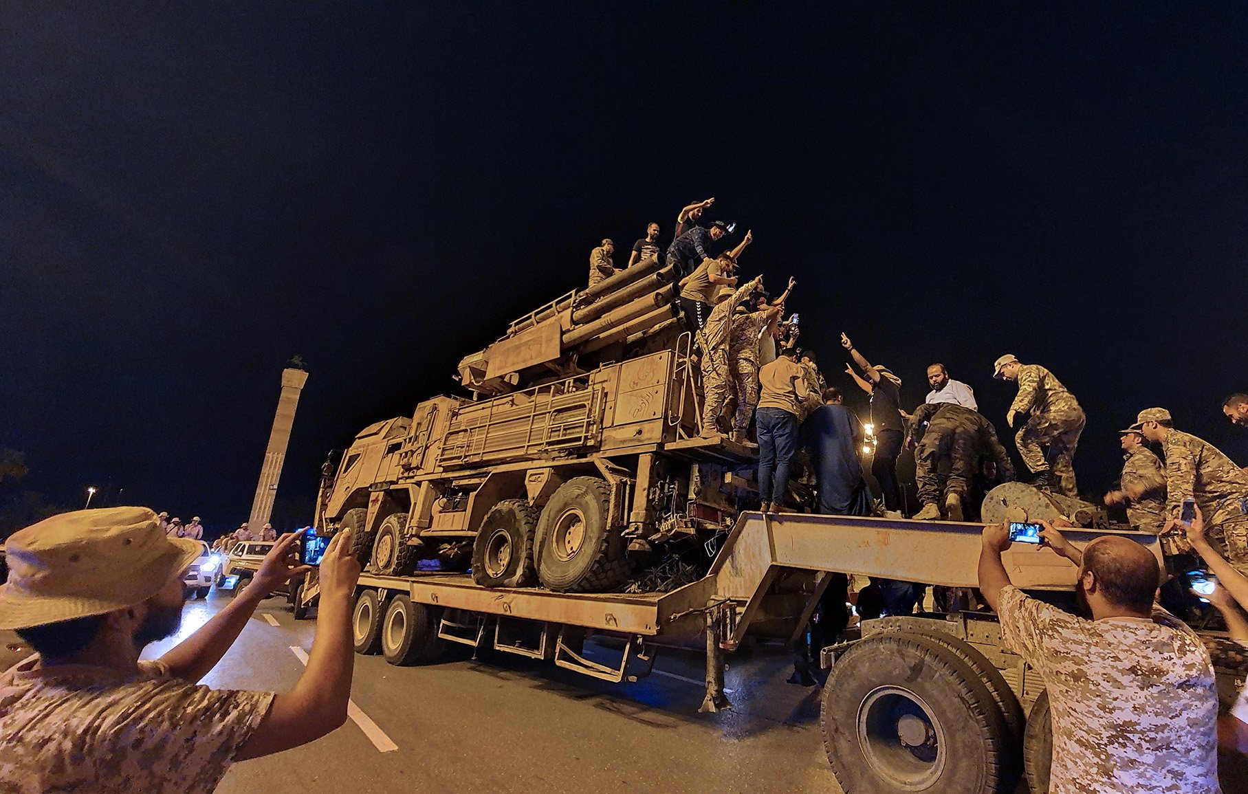 L'immagine mostra un gruppo di persone radunate intorno a un veicolo militare, che sembra essere un camion con un modulo di artiglieria. La scena si svolge di notte, con luci artificiali che illuminano l'ambiente. Alcuni individui sono saliti sopra il veicolo, mentre altri stanno scattando foto con i loro telefoni. L'atmosfera sembra carica di emozione e partecipazione, con gesti di saluto o di celebrazione da parte delle persone sopra il veicolo.