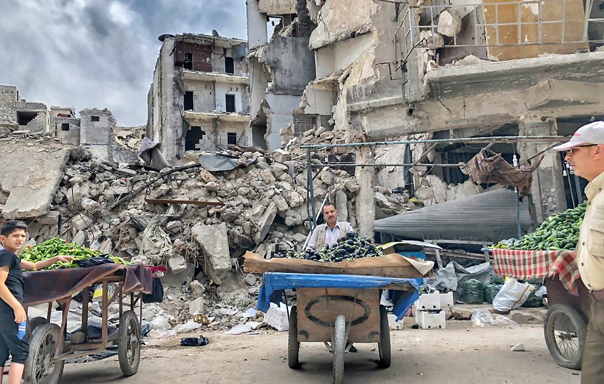 L'image montre une scène d'un marché ou d'une vente de légumes dans un environnement dévasté, avec des bâtiments en ruine en arrière-plan. On voit deux personnes, probablement des vendeurs, avec des charrettes remplies de légumes, comme des courgettes ou des poivrons. L'atmosphère est chargée, avec des nuages obscurcis qui semblent se rassembler, ajoutant à la gravité de la situation. Les décombres et les bâtiments effondrés indiquent une zone touchée par un conflit ou une catastrophe.