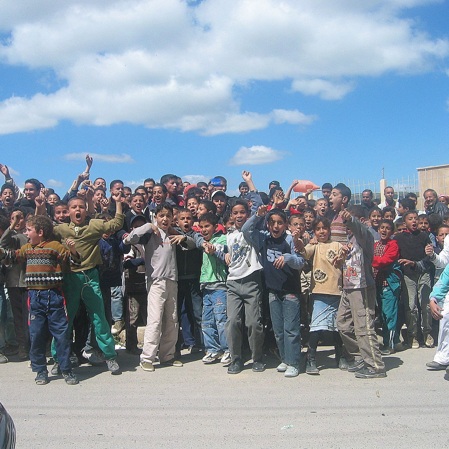 L'image montre un groupe important d'enfants et d'adolescents rassemblés en plein air. Ils semblent joyeux et excités, levant les bras et faisant des gestes. Le ciel est clair avec quelques nuages, et l'environnement urbain en arrière-plan suggère un cadre scolaire ou communautaire. Les enfants portent des vêtements variés, reflet de leur diversité. L'atmosphère semble animée et festive.