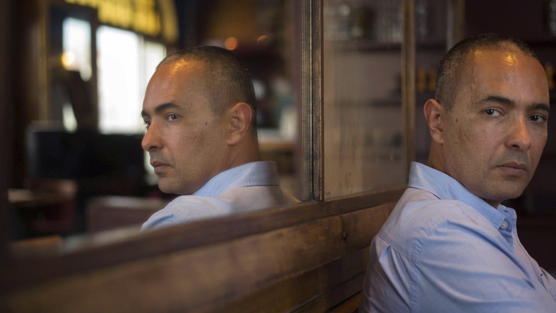 The image features a man sitting with his back to a reflective surface, creating a mirror effect. He has short, dark hair and is wearing a light blue shirt. His expression is pensive, with a serious look on his face as he gazes thoughtfully to the side. The surroundings indicate a cozy indoor setting, with warm lighting and wooden elements in the background. The reflection captures both his profile and the contemplative mood of the scene.
