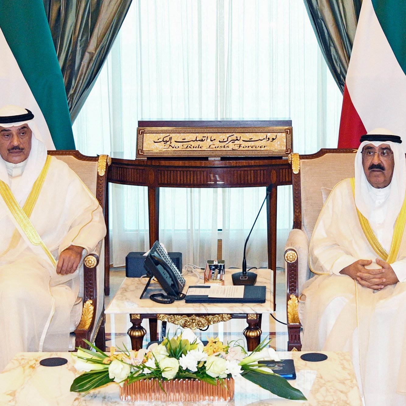 L'image montre deux hommes assis dans une salle de réunion élégante. Ils portent des vêtements traditionnels, possiblement des habitudes de type arabe, avec des écharpes sur la tête. Leurs expressions sont sérieuses et ils sont entourés de décorations luxueuses, comme des rideaux drapés et des meubles en bois. Au centre, il y a une table en marbre avec quelques objets, et en arrière-plan, on peut voir un tableau avec des inscriptions arabes. L'atmosphère dégage un sentiment de formalité et de respect.