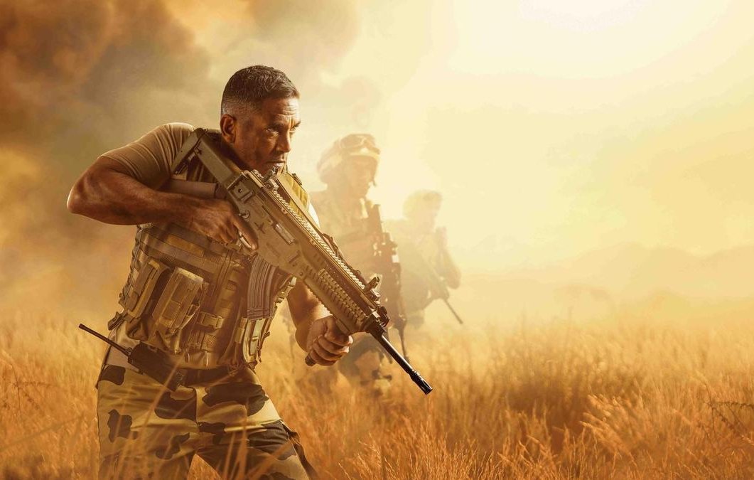 L'image montre des soldats en pleine opération militaire, évoluant dans un environnement désertique. Un homme au premier plan est en position de tir, portant un uniforme de camouflage et un gilet tactique. Il semble concentré et déterminé. En arrière-plan, d'autres soldats avancent, créant une atmosphère de tension et d'action. La lumière est chaude, avec des teintes dorées, suggérant un moment critique dans un paysage aride.