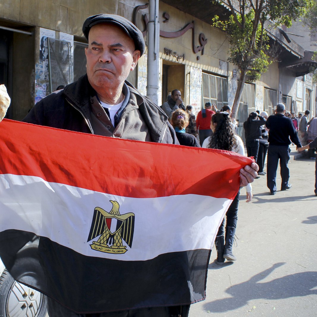 L'image montre un homme tenant un drapeau égyptien, avec le symbole de l'Égypte au centre. Il tient également un morceau de pain dans l'autre main. Demain la ville est animée, avec plusieurs personnes en arrière-plan, ce qui suggère une atmosphère de rassemblement ou de manifestation. L'homme semble sérieux et déterminé, exprimant un fort sentiment national. L'environnement urbain, avec des bâtiments et des passants, reflète la vie quotidienne en Égypte.