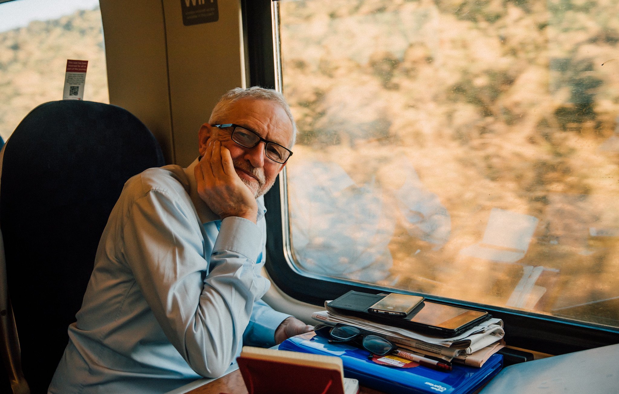 L'image montre un homme assis à côté d'une fenêtre de train. Il porte une chemise claire et a des lunettes. Son visage exprime une certaine réflexion ou contemplation alors qu'il repose sa tête sur sa main. Sur la table devant lui, on peut voir plusieurs objets comme des téléphones, des lunettes de soleil et des papiers, donnant l'impression qu'il a passé du temps à travailler ou à organiser ses affaires. À l'extérieur de la fenêtre, on aperçoit un paysage flou, suggérant que le train est en mouvement.