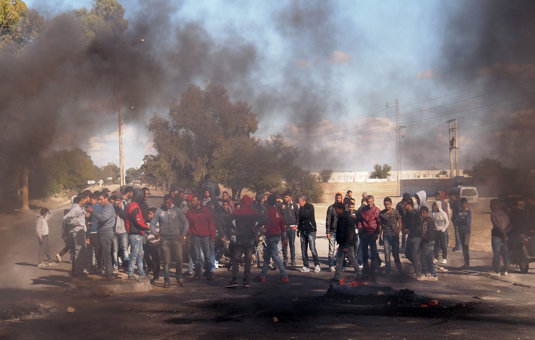 L'image montre un groupe de personnes rassemblées sur une route où de la fumée noire s'élève, probablement à cause de pneus ou d'autres matériaux en feu. Les manifestants semblent préoccupés et engagés dans un rassemblement, tandis que l'arrière-plan indique un paysage urbain avec des arbres et des infrastructures. La scène évoque une atmosphère de tension ou de protestation.