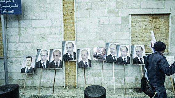 L'image montre un mur en béton avec plusieurs portraits accrochés, principalement des visages d'un homme en costume. Ces portraits sont placés sur des supports en bois. Une personne, dont on ne voit que le dos, marche à proximité. Le décor semble urbain et pourrait impliquer un contexte politique. Les expressions faciales sur les portraits sont sérieuses, reflétant une ambiance de propagande ou de soutien.