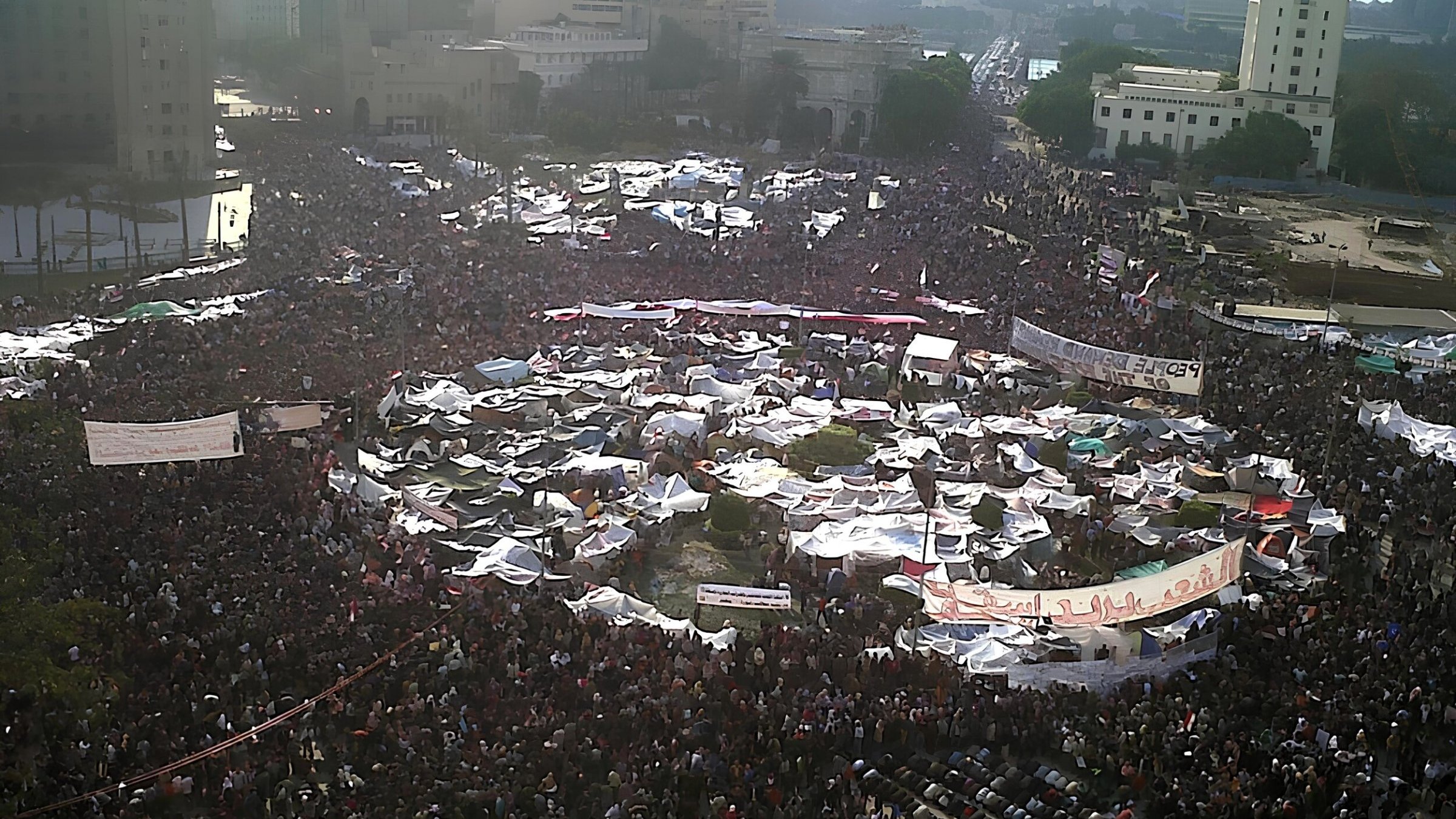 Una grande folla si raduna in una piazza, con tende e striscioni.