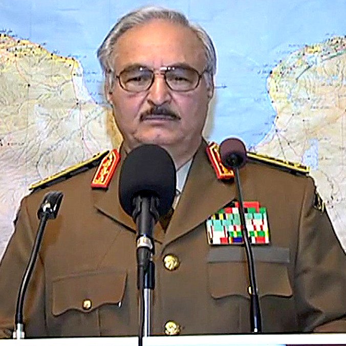 L'image montre un homme portant un uniforme militaire, avec plusieurs médailles et distinctions sur sa veste. Il se tient derrière un pupitre de conférence, devant une carte géographique en arrière-plan. L'homme semble s'adresser à un public ou à une caméra. On peut voir des drapeaux en arrière-plan, indiquant une situation officielle.