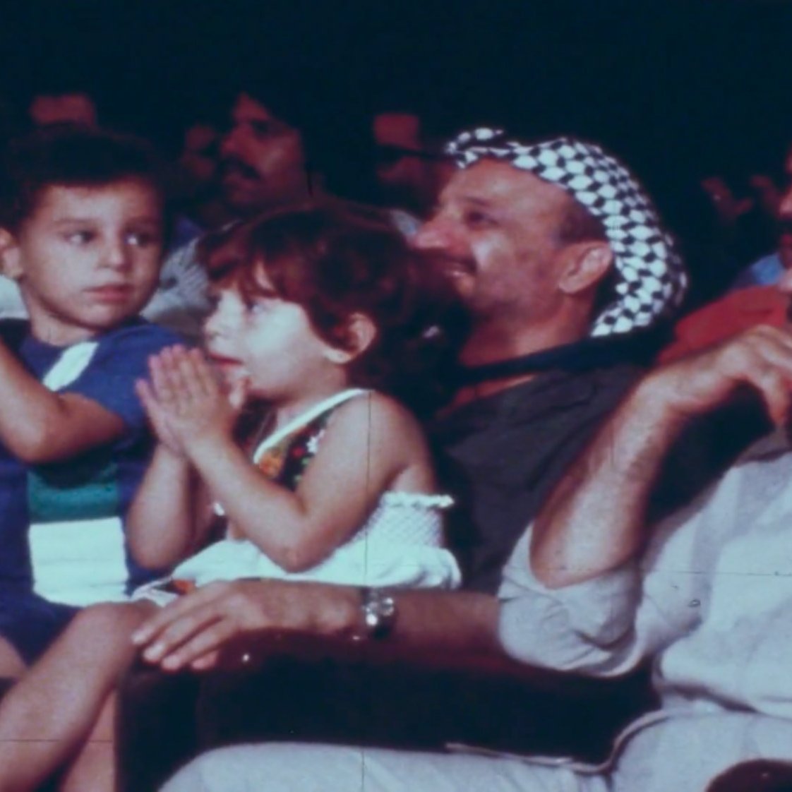 في الصورة مجموعة من الأشخاص يجلسون في قاعة، ومن بينهم طفلان يجلسان أمام رجل يرتدي كوفية. يبدو أن الجميع يستمع أو يشاهد شيئًا ما في الفعالية. الأطفال يظهرون تفاعلهم بالتصفيق أو الاستمتاع بالعرض. الرجال في الصورة يبدون مهتمين ومسترخين. الجو يبدو احتفاليًا أو حيويًا.