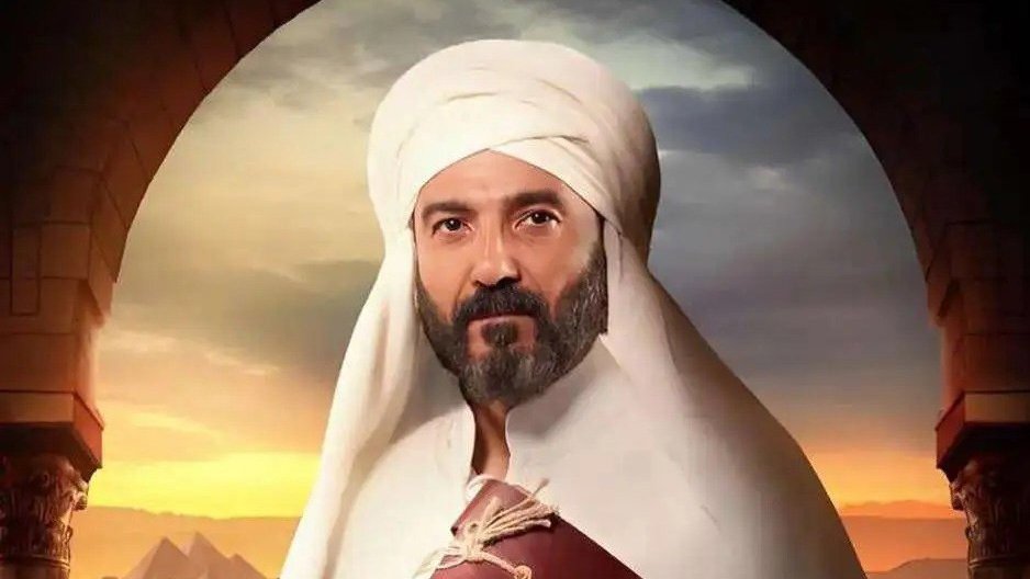L'image représente un homme portant un turban et une tenue traditionnelle. Il a une barbe et une expression sérieuse. En arrière-plan, on aperçoit un paysage désertique avec des pyramides, suggérant une ambiance historique ou culturelle. La lumière semble douce, ajoutant une atmosphère dramatique à la scène.