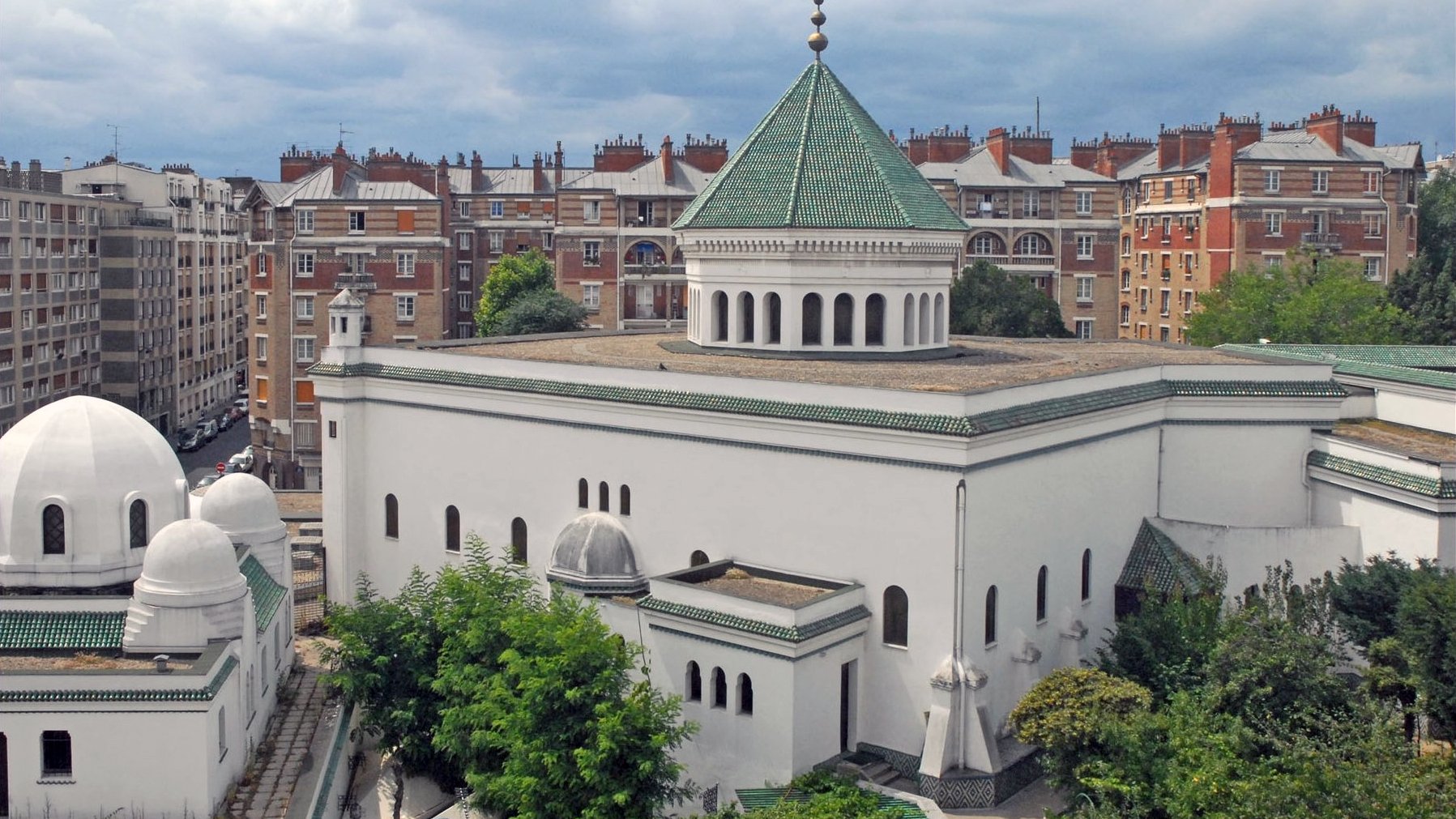 L'image montre un bâtiment architectural distinctif avec une façade blanche et un toit en tuiles vertes. Le design présente des éléments inspirés de l'architecture islamique, comme des dômes et des arches. En arrière-plan, on aperçoit des immeubles de style haussmannien, typiques de nombreuses villes françaises, avec des balcons et des toits en attique. Le ciel est nuageux, ce qui ajoute une atmosphère particulière à la scène. Des arbres et de la verdure entourent le bâtiment, contribuant à l'esthétique générale.