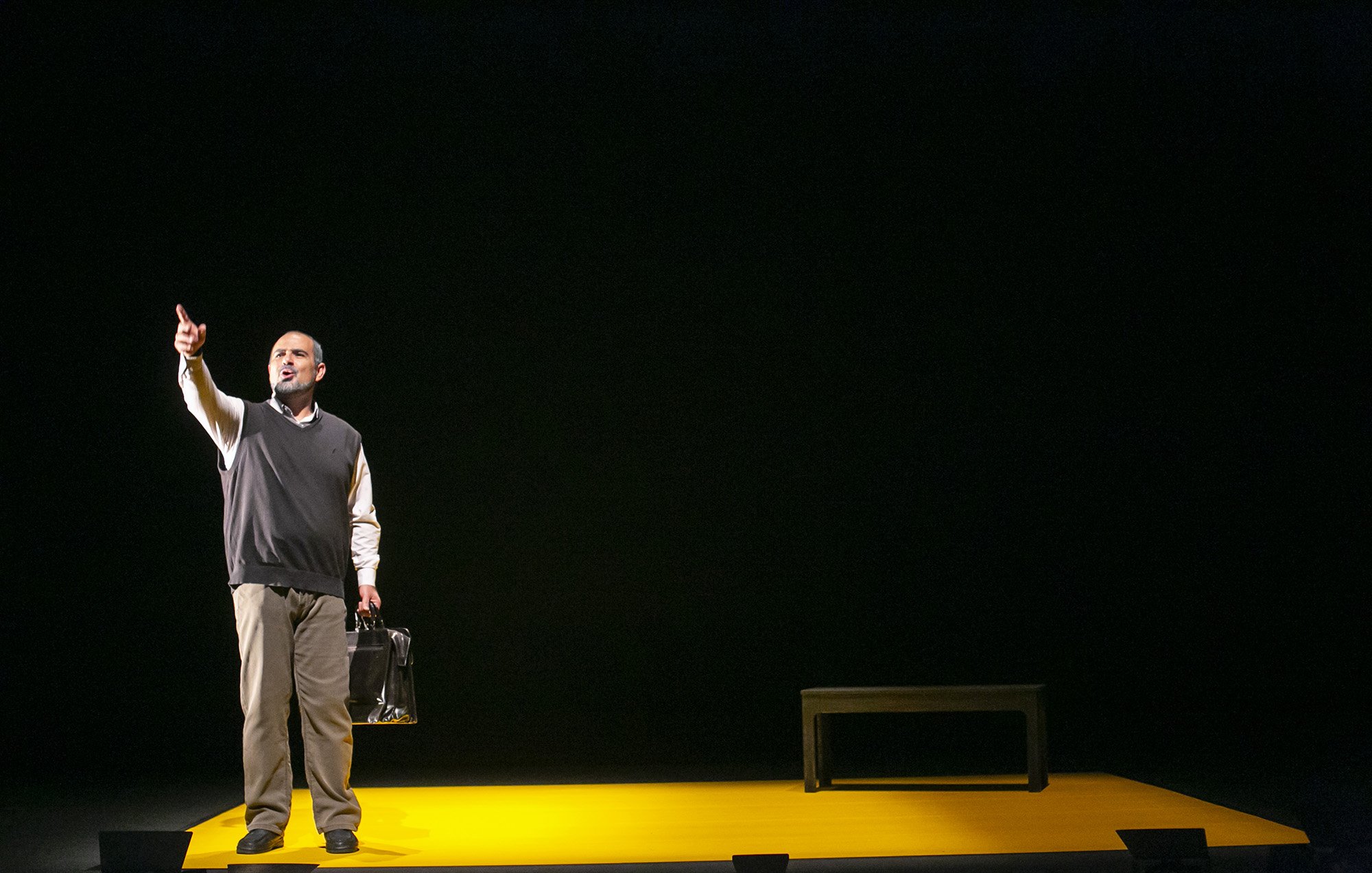 L'image montre un homme sur scène, debout sur un tapis jaune. Il porte un pull noir par-dessus une chemise blanche et tient une serviette ou un sac à main à la main. L'homme semble pointer vers le haut, exprimant une émotion forte ou un message. La scène est sombre autour de lui, ce qui met l'accent sur sa présence au centre et sur l'objet noir qui pourrait être un banc ou une table. L'ambiance donne une impression de sérieux ou de réflexion.