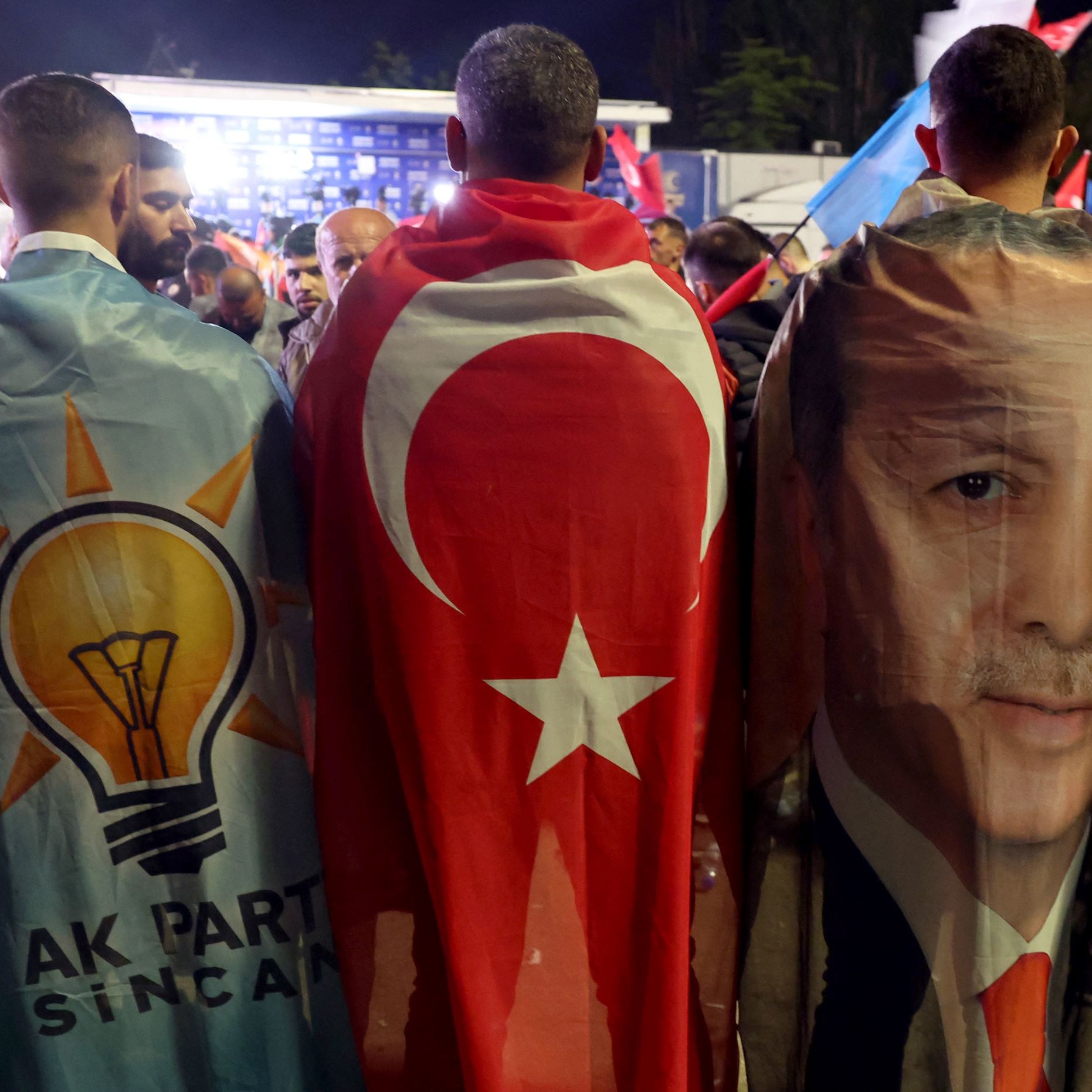 L'image montre un groupe de personnes réunies lors d'un événement politique, enveloppées dans des drapeaux et des bannières. Deux d'entre elles portent des capes : l'une présente le symbole du parti AKP (Parti de la justice et du développement), tandis que l'autre arbore le drapeau turc et une image du président Erdoğan. On peut voir une ambiance de rassemblement avec des drapeaux turcs en arrière-plan, suggérant une atmosphère de soutien et de célébration.