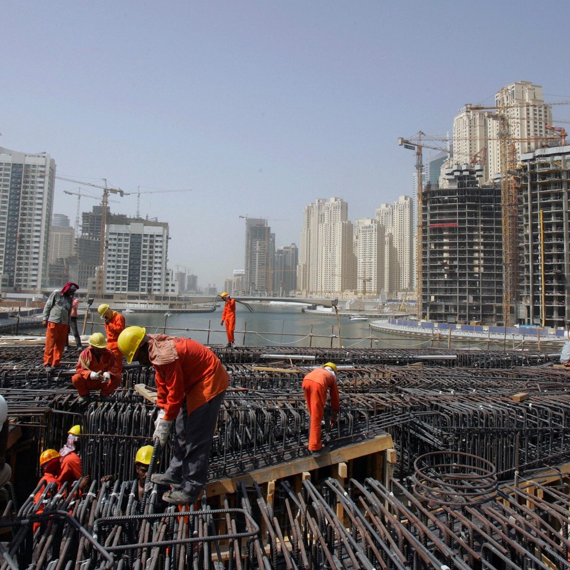 الصورة تظهر موقع بناء في منطقة حضرية حديثة. يتواجد فيها العديد من العمال يرتدون ملابس العمل البرتقالية والخوذ البيضاء. تشير الصورة إلى أن العمال مشغولون بعمليات البناء، حيث يمكن رؤية الهياكل الحديدية والرافعات في الخلفية. المباني الشاهقة الحديثة المحيطة بالموقع تدل على تطور المدينة، مع وجود بعض العمارات قيد الإنشاء. السماء تبدو صافية، ولكن هناك غبار خفيف يوحي بنشاط البناء.