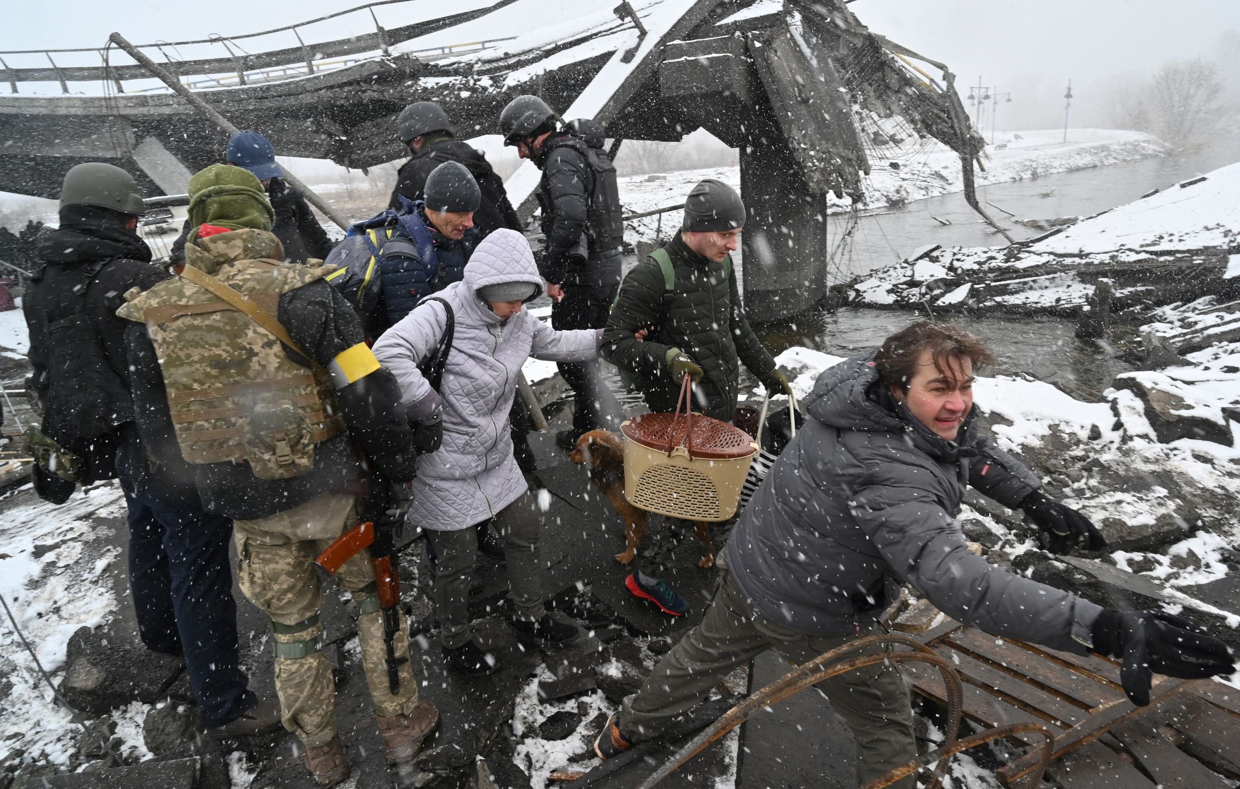 L'immagine mostra un gruppo di persone in un'area distrutta, probabilmente a causa di un conflitto o di un disastro. Alcune persone indossano abbigliamento militare, mentre altre sembrano civili. È in corso un salvataggio o un'evacuazione, con qualcuno che sta aiutando un'altra persona a attraversare il terreno difficile. La scena è caratterizzata da neve e condizioni atmosferiche difficili, suggerendo un ambiente critico e di emergenza. I volti mostrano preoccupazione e determinazione.