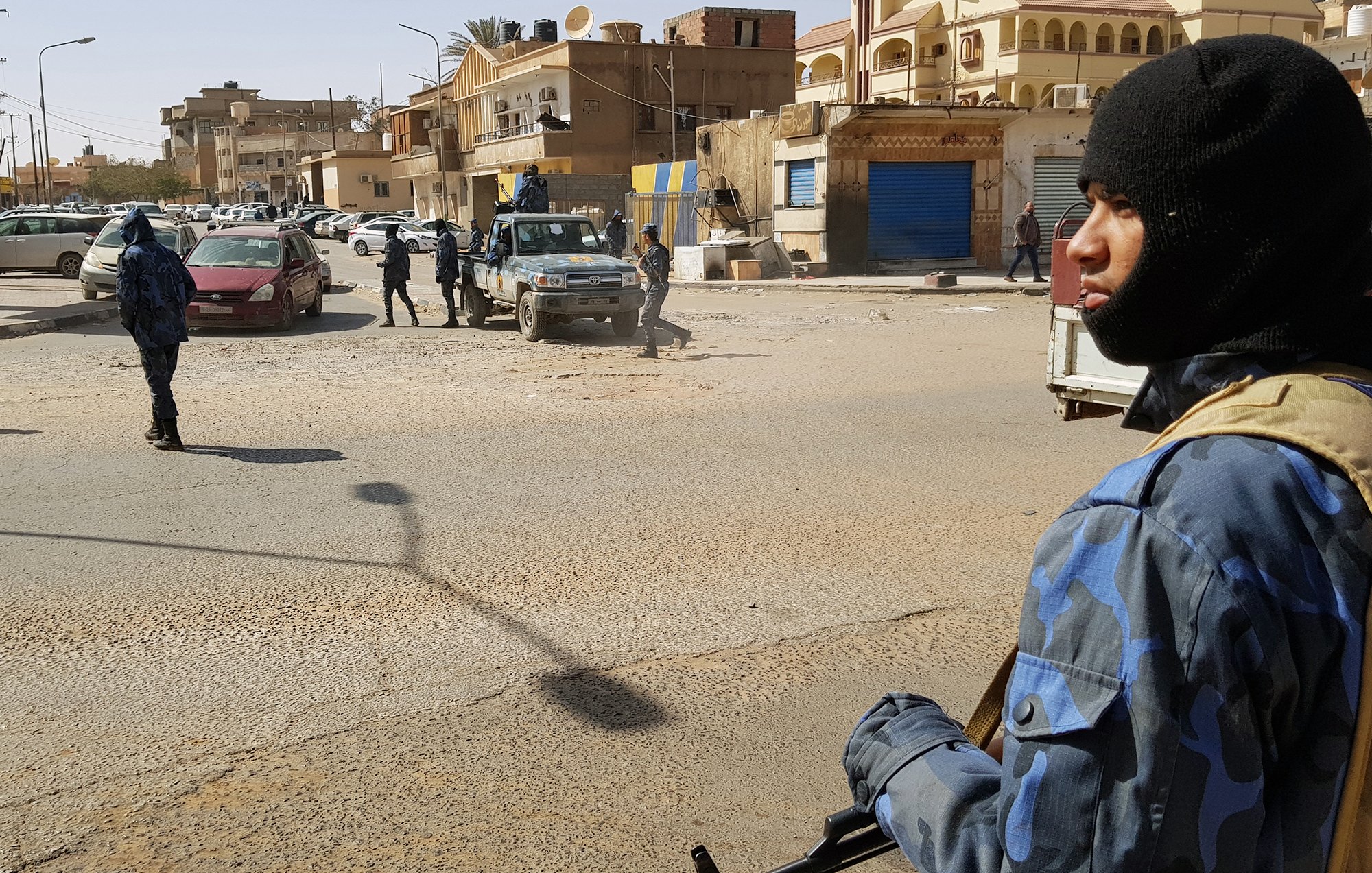 تظهر الصورة مشهداً في منطقة حضرية، حيث يوجد عدد من رجال الأمن يرتدون زيًّا رسميًّا وقبعات سُوداء. أحدهم يحمل سلاحًا ويقف في المقدمة، بينما يتواجد آخرون في الخلفية. تشهد المنطقة حركة مرورية، مع وجود سيارات متوقفة في الشارع. المباني المحيطة تظهر بألوان متنوعة، وبعضها يبدو أنه مغلق. تعكس الصورة أجواءً من اليقظة والحذر الأمني.