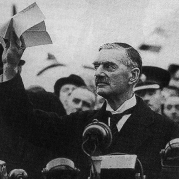 L'image montre un homme en costume, se tenant devant un microphone avec une expression grave. Il lève un ou plusieurs documents au-dessus de sa tête, visiblement en train de s'adresser à une foule. En arrière-plan, on peut voir des personnes assemblées, portant des uniformes et écoutant attentivement. L'atmosphère semble être celle d'un discours important, probablement lié à des événements politiques.