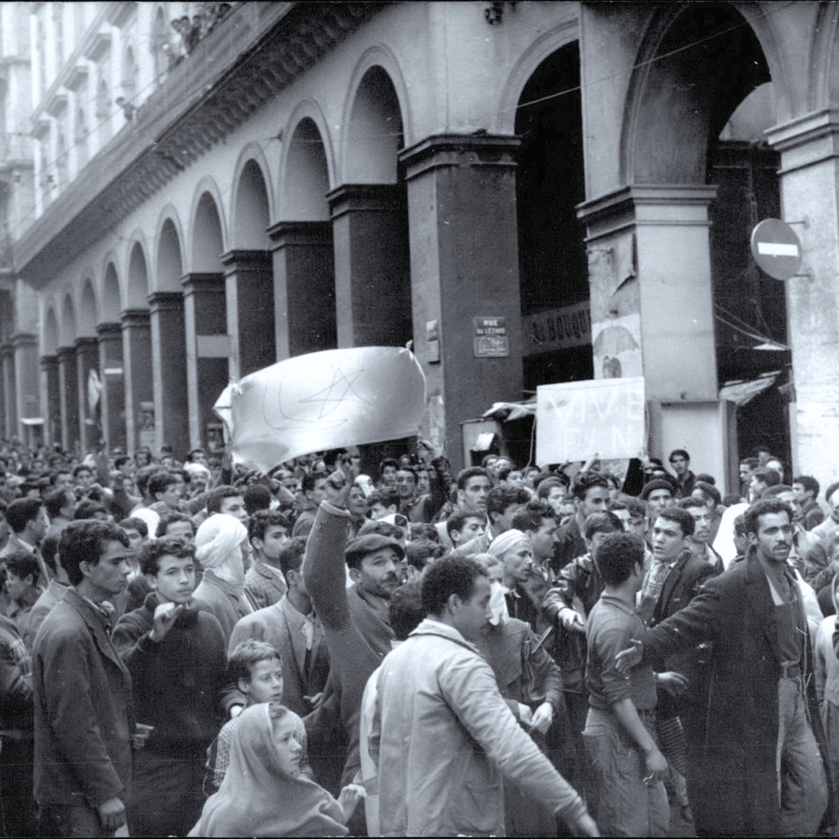 L'image montre une grande manifestation dans une rue animée, où une foule dense est rassemblée. Les manifestants semblent exprimer des revendications, brandissant des drapeaux et des pancartes. L'architecture environnante est typiquement urbaine, avec de grands arcs et des bâtiments anciens. L'atmosphère générale semble passionnée et engagée, reflétant un fort esprit de protestation.