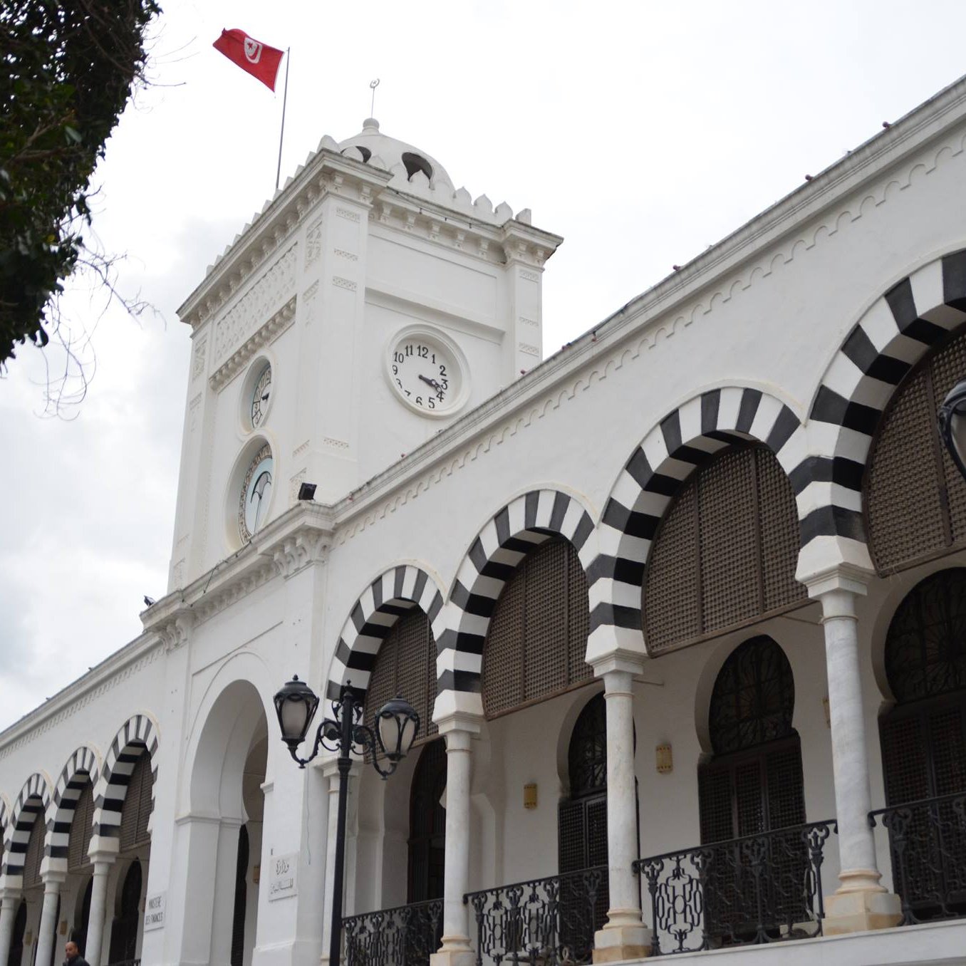 L'image montre un bâtiment architectural blanc avec des arcades noires et blanches. Il y a une horloge sur la façade et un drapeau tunisien flottant au sommet. Le ciel est nuageux, ce qui donne une ambiance légèrement sombre. Des lampadaires sont visibles le long de la structure, ajoutant une touche d'élégance à l'ensemble.
