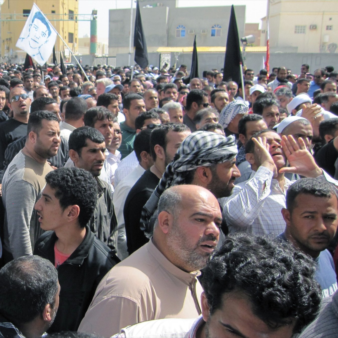 L'image montre une grande foule de personnes rassemblées, probablement lors d'une manifestation. Les participants semblent très engagés, certains tenant des drapeaux, y compris celui de Bahreïn. Les visages expriment diverses émotions, allant de la détermination à l'interrogation. L'environnement montre des bâtiments en arrière-plan, suggérant une zone urbaine. L'atmosphère générale dénote une forte implication politique ou sociale.