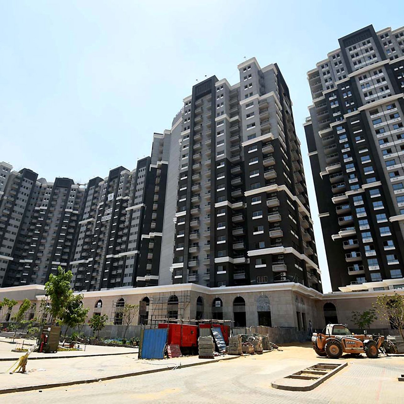 L'image montre un ensemble de grands immeubles résidentiels modernes. Les bâtiments sont principalement de couleur noire et blanche, avec de grandes fenêtres qui laissent entrer la lumière. On peut voir un chantier en cours au premier plan, avec des véhicules de construction et des matériaux dispersés. Il y a aussi quelques arbres et des espaces verts, suggérant un aménagement paysager en développement. Le ciel est dégagé et ensoleillé.