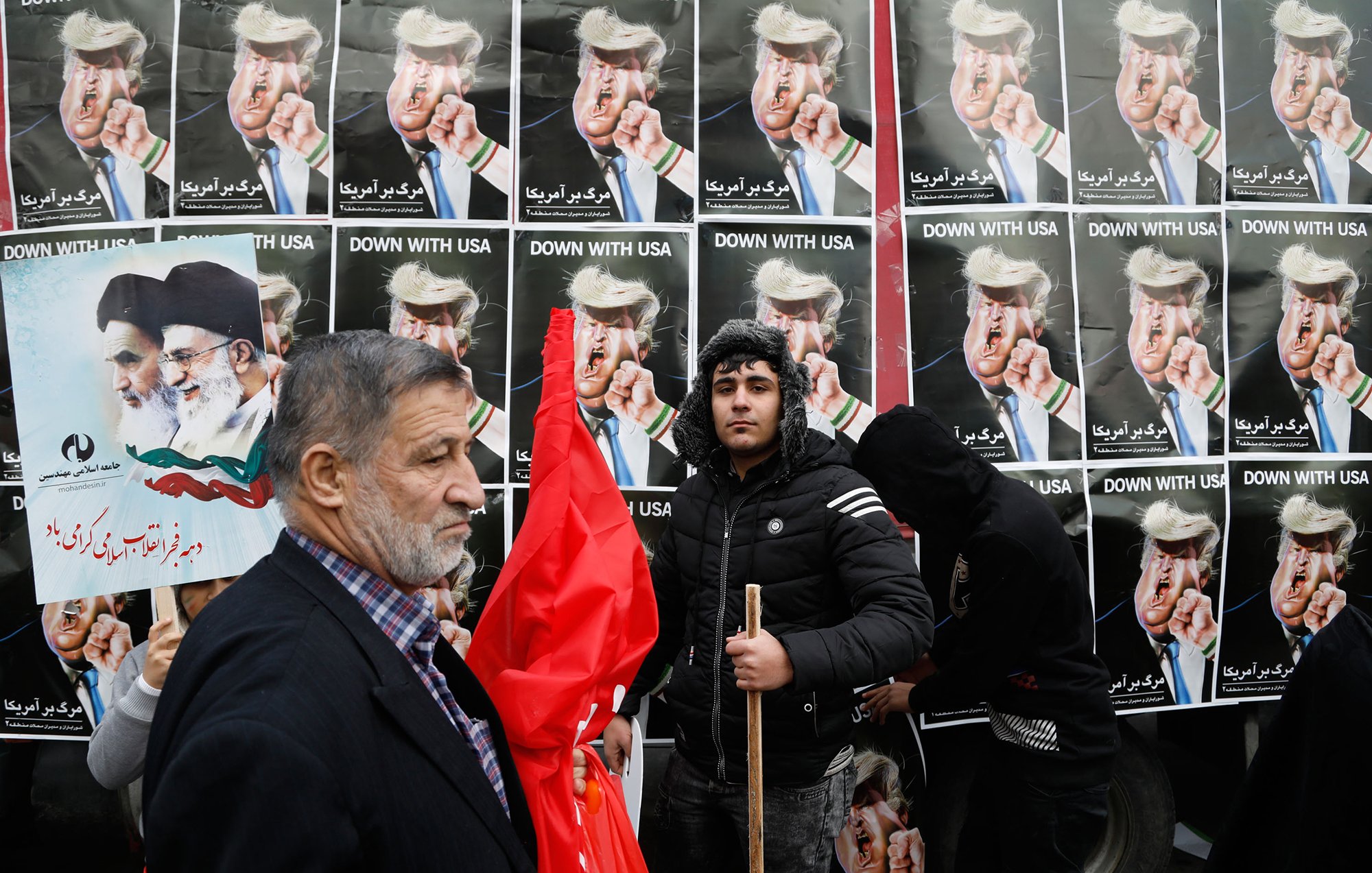 L'image montre une manifestation avec des affiches en arrière-plan affichant des personnages aux visages provocateurs, probablement des figures politiques, portant des coiffures spécifiques. Des gens tiennent des pancartes et des drapeaux, tandis qu'un homme sur le devant semble exprimer un message clair de désaccord ou de protestation. L'ensemble de la scène dégage une ambiance de contestation, soulignant des tensions politiques.