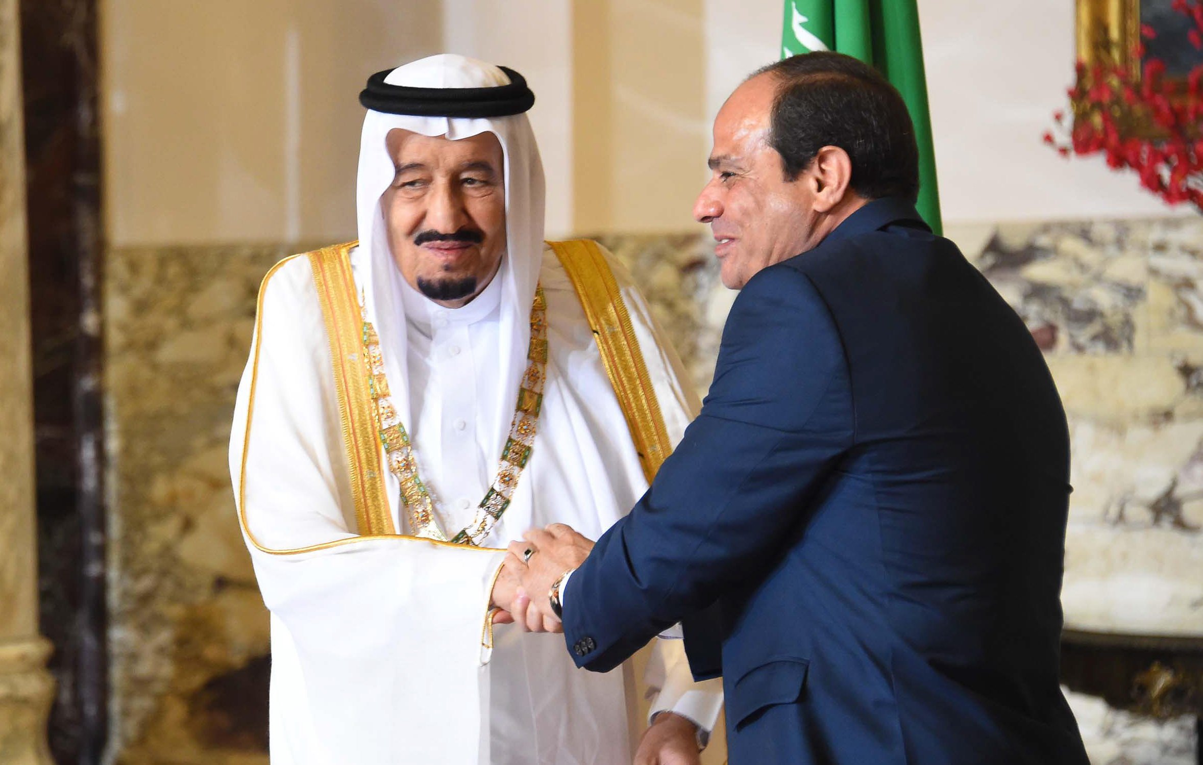 L'image montre deux hommes en costume, se serrant la main dans un cadre formel. L'un est vêtu d'une robe traditionnelle saoudienne et d'un couffin, tandis que l'autre porte un costume sombre. Ils semblent échanger un sourire, ce qui indique une interaction amicale. En arrière-plan, on peut apercevoir des décorations et des drapeaux, suggérant un contexte diplomatique ou officiel.