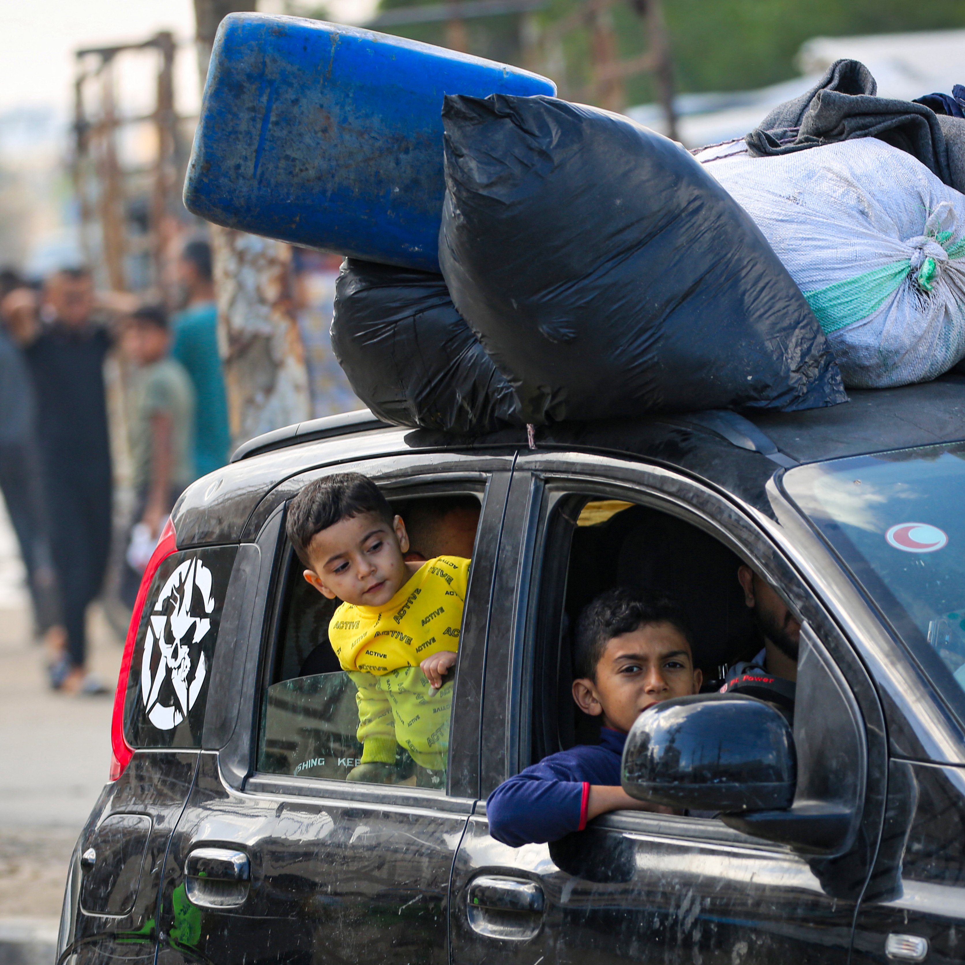 L'immagine mostra un'auto con un carico sopra di sacchi neri e altri oggetti. All'interno della vettura, si vedono due bambini: uno che guarda fuori dal finestrino con espressione curiosa e l'altro che si affaccia dall'apertura. Sullo sfondo si possono notare altre persone e un ambiente urbano, che sembra un'area affollata. La scena riflette una situazione di vita quotidiana in un contesto urbano.