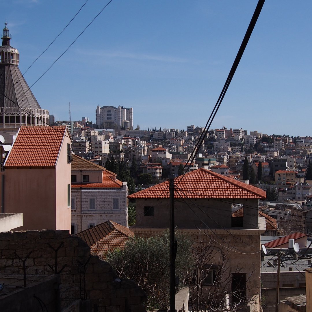 L'image montre un paysage urbain avec des bâtiments aux toits en tuiles rouges, typiques de certaines architectures méditerranéennes. On peut observer un dôme sur l'un des bâtiments, sans doute une église ou un édifice religieux. En arrière-plan, la ville s'étend sur plusieurs niveaux, avec de nombreuses constructions. Le ciel est dégagé et ensoleillé, créant une ambiance lumineuse. Des lignes électriques traversent l'image, ajoutant un élément urbain au paysage.
