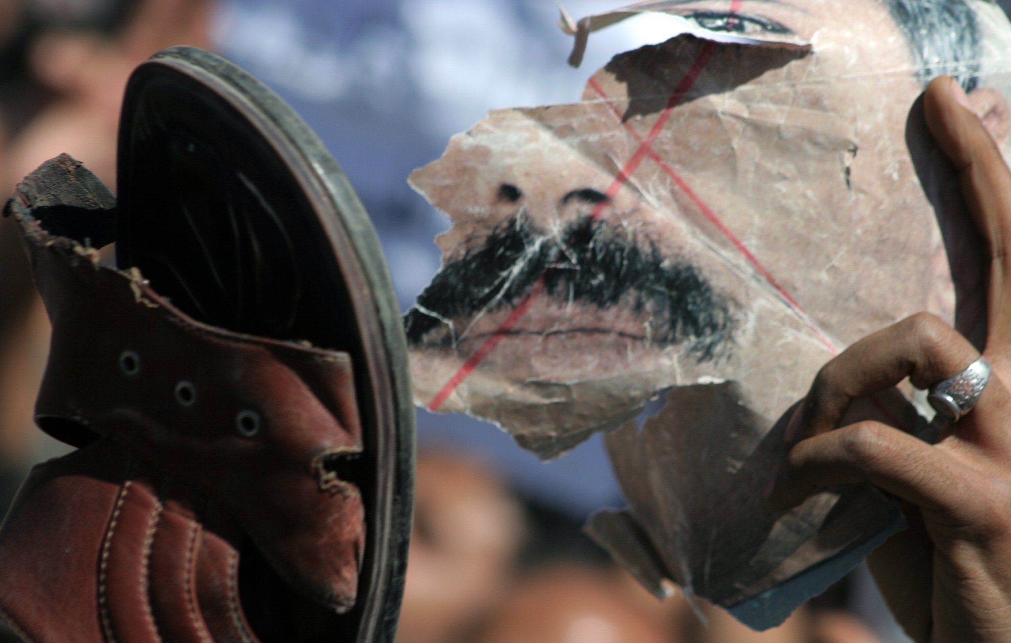 L'image montre une chaussure levée vers un visage déchiré, probablement représentant une personne. Le visage est marqué par des lignes rouges et semble avoir été martelé ou abîmé. L'atmosphère de l'image suggère de la contestation ou de l'opposition, avec une foule floue en arrière-plan. La chaussure et le visage déchiré symbolisent peut-être un acte de rébellion ou de rejet.