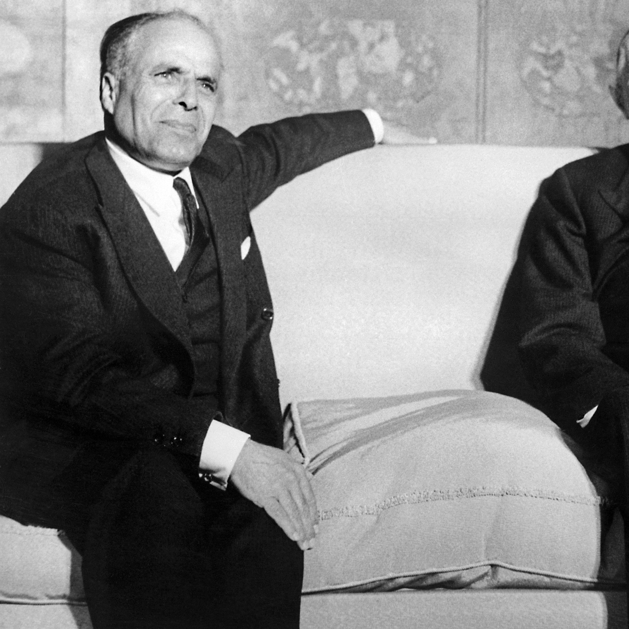 L'image montre deux hommes assis sur un canapé dans un environnement formel. Ils ont l'air sérieux et portent des costumes sombres. L'un des hommes semble légèrement se pencher en arrière, tandis que l'autre regarde devant lui. L'arrière-plan est décoré, mais les détails ne sont pas clairement visibles. L'atmosphère générale apparaît académique et solennelle.