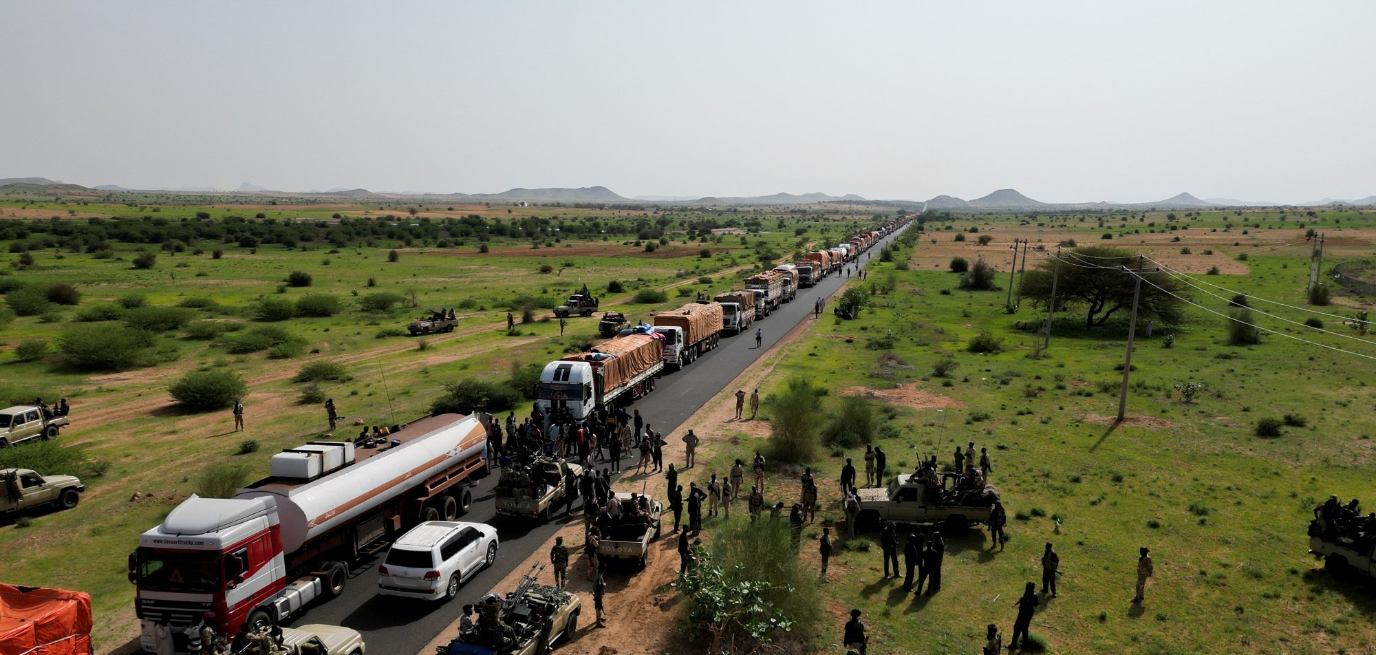 A long line of trucks on a road, with soldiers and green fields surrounding them.