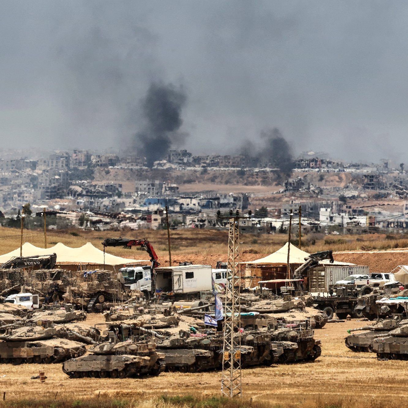 Un champ militaire avec des chars, fumée au loin et destructions visibles en arrière-plan.