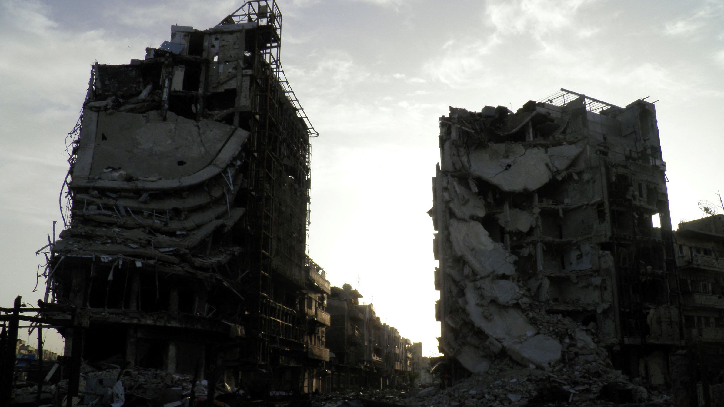 L'image montre deux bâtiments en ruines, témoignant des conséquences d'un conflit ou d'une catastrophe. Les structures sont partiellement effondrées, avec des débris dispersés autour. L'éclairage est sombre, suggérant un crépuscule ou une journée nuageuse, ce qui ajoute une atmosphère de désolation à la scène. Le paysage environnant semble aussi dévasté, accentuant la gravité de la situation.