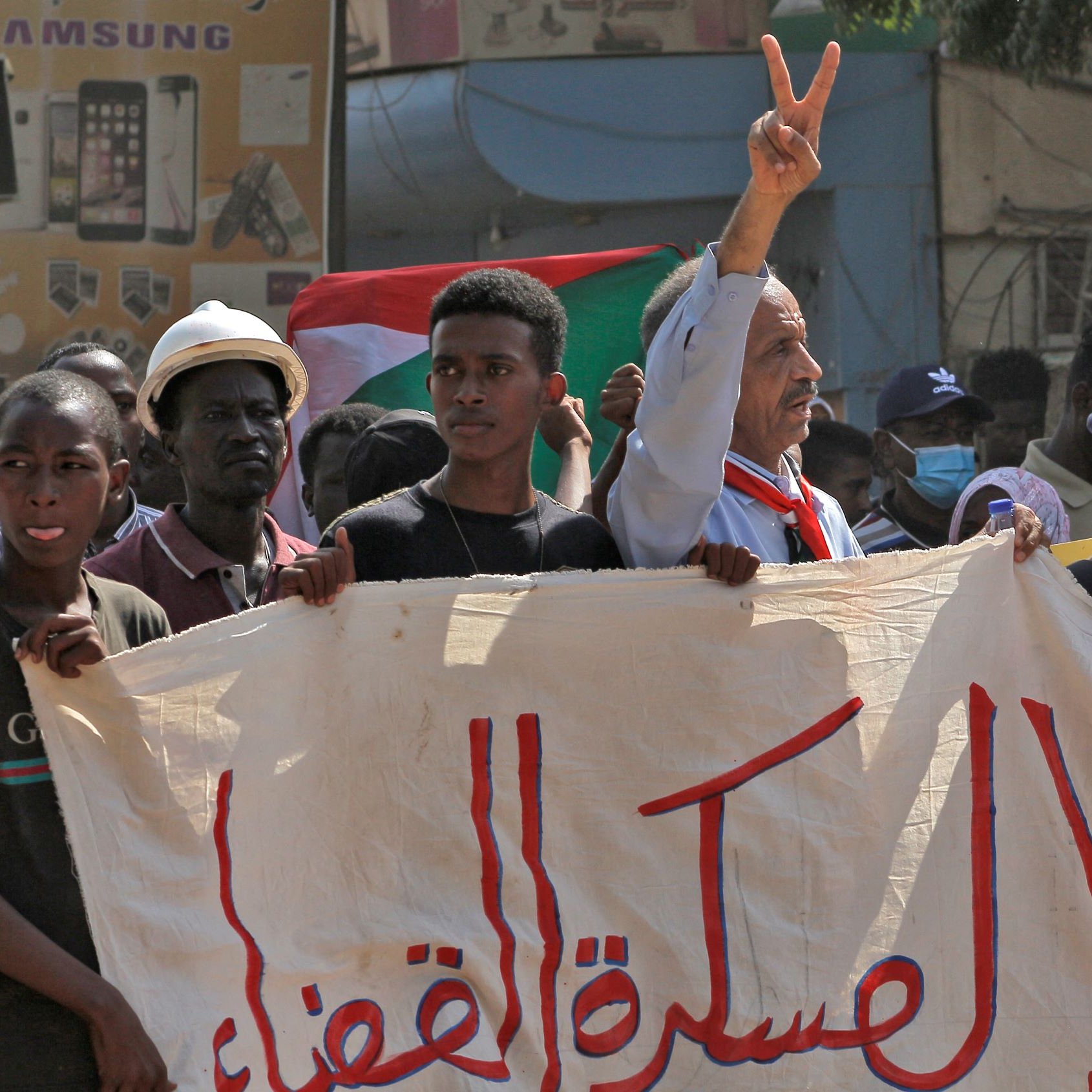 L'immagine mostra una manifestazione con diverse persone che sollevano cartelli e bandiere. In primo piano, un uomo con un cappello e una sciarpa al collo alza la mano mostrando il simbolo della pace. Gli altri manifestanti sembrano esprimere supporto per una causa, cominciando da una grande bandiera sudanese. Lo sfondo mostra dei cartelloni pubblicitari, mentre l'atmosfera appare carica di energia e determinazione.