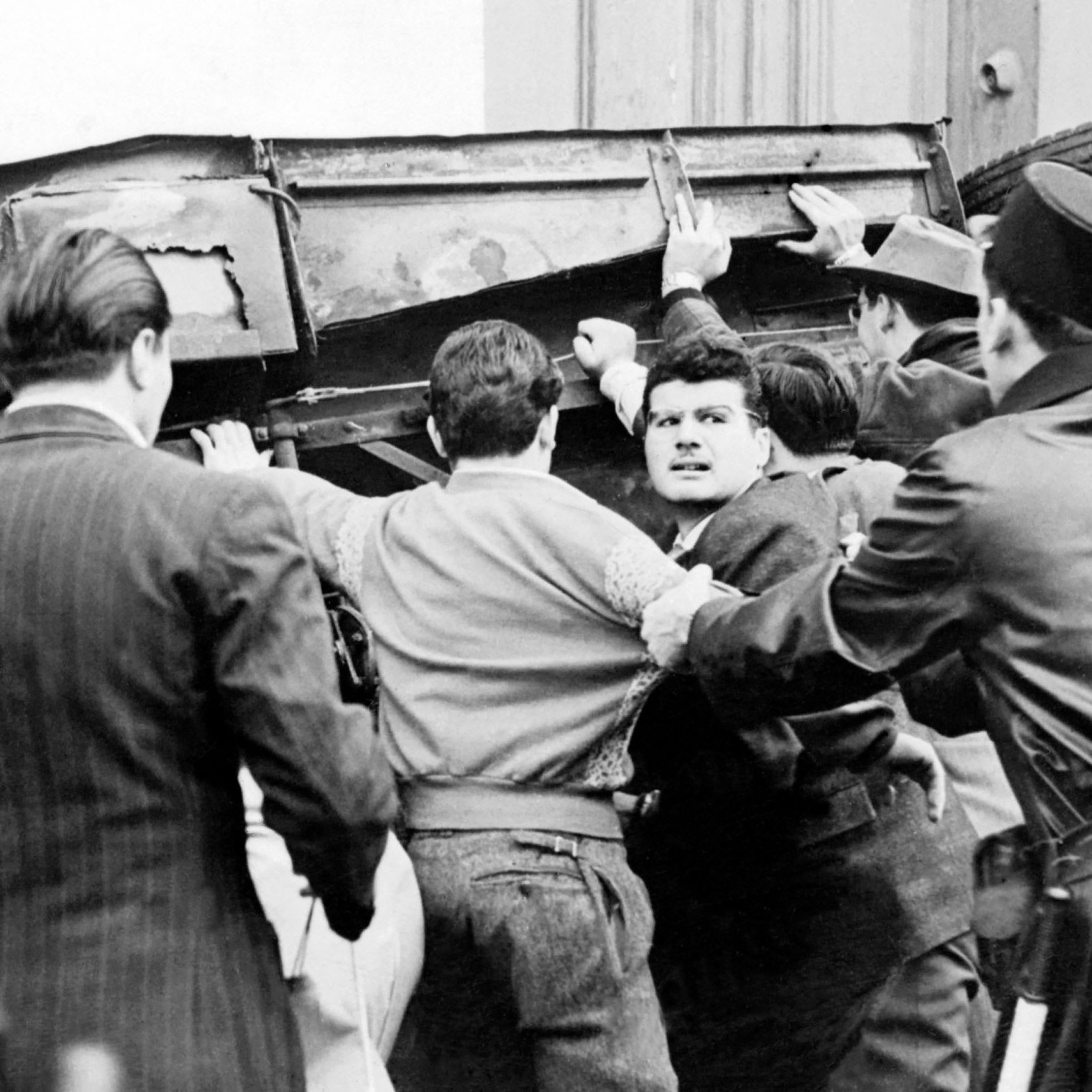 L'image montre une scène tumultueuse où un groupe de personnes semble être impliqué dans une altercation. Au centre, plusieurs hommes s'efforcent de soulever un véhicule renversé, tandis qu'un homme au visage concentré semble être en train de s'opposer à la situation ou d'interagir avec les autres. À droite, un policier en uniforme semble gérer la situation. L'atmosphère est tendue, et le contraste en noir et blanc accentue l'intensité de la scène.