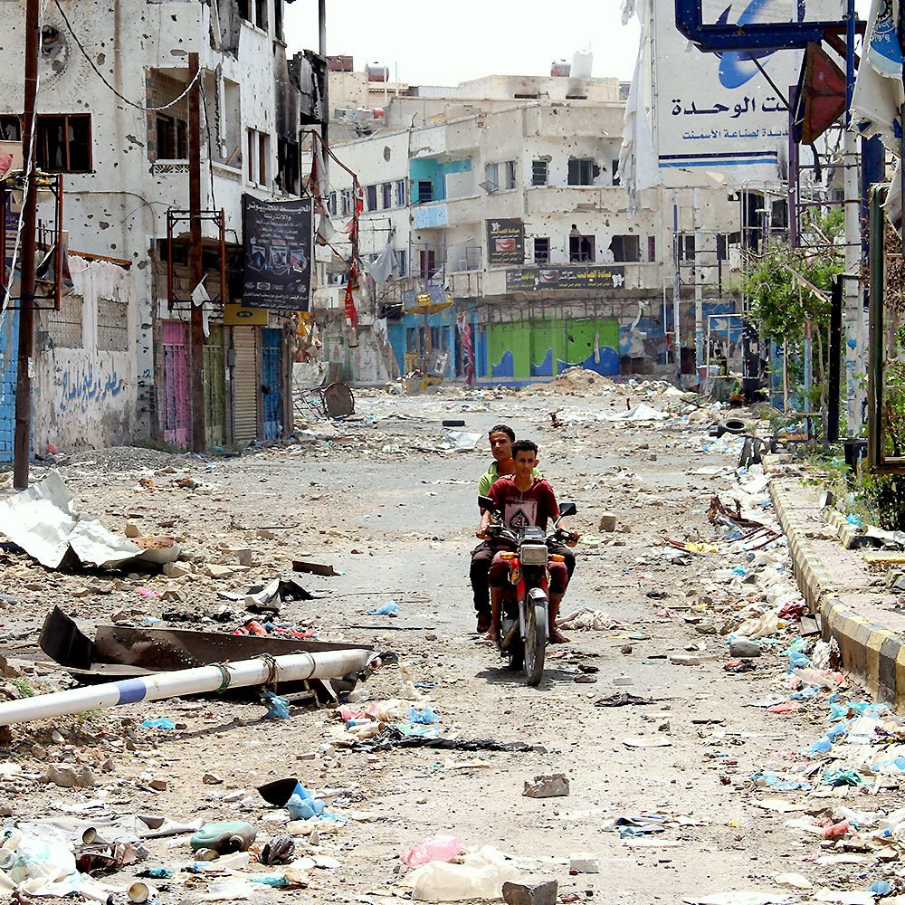 L'image montre une scène de rue désolée et abandonnée, probablement dans une zone urbaine touchée par des conflits ou des désastres. Le sol est recouvert de débris et de détritus, tandis que les bâtiments autour semblent endommagés. Au centre de l'image, deux personnes sont assises sur une moto, traversant cette rue dévastée. Les enseignes et les murs portent des marques de détérioration, reflétant un environnement difficile et chaotique.