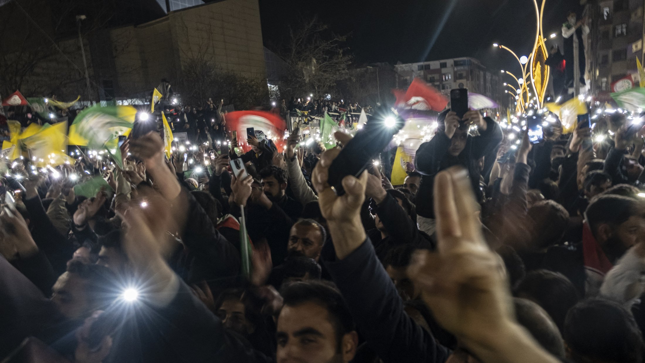 L'image montre une grande foule rassemblée dans un lieu public, probablement lors d'une manifestation ou d'une célébration nocturne. Les participants brandissent des drapeaux colorés et lèvent les mains, certains tenant des téléphones allumés qui éclairent la scène. L'atmosphère semble festive et énergique, avec des expressions de joie et d'engagement visible sur les visages des gens. Les bâtiments environnants sont sombres, accentuant les lumières des téléphones et des drapeaux.
