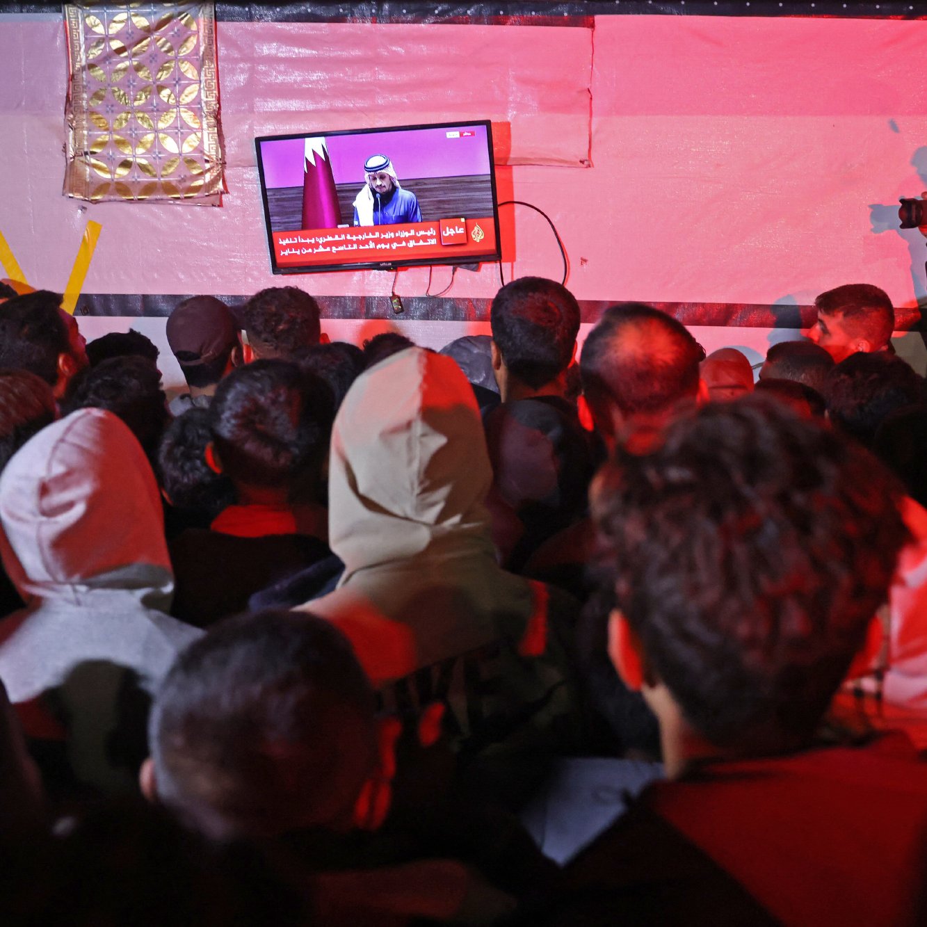 L'image montre une foule de personnes rassemblées, observant attentivement un écran au-dessus d'eux. L'écran affiche un discours ou une annonce, probablement en rapport avec des événements d'actualité. Certaines personnes dans la foule portent des capuches et des vêtements chauds, ce qui suggère qu'il fait frais. Un individu à droite prend des photos, ajoutant une dimension journalistique à la scène. L'éclairage est tamisé, créant une ambiance particulière.