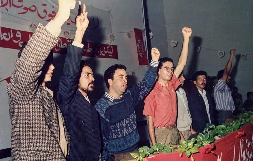 تظهر الصورة مجموعة من الأشخاص واقفين خلف منصة، يرفعون أيديهم في إشارة للتعبير عن الاحتجاج أو الدعم. الخلفية تحتوي على لافتات مكتوبة، مما يشير إلى أن الحدث قد يكون سياسيًا أو اجتماعيًا. الجو العام يبدو حماسيًا، حيث يعكس تفاعل الجمهور مع ما يحدث أمامهم. الأشخاص يرتدون ملابس تتنوع في الألوان والأساليب، مما يعكس حقبة زمنية معينة.