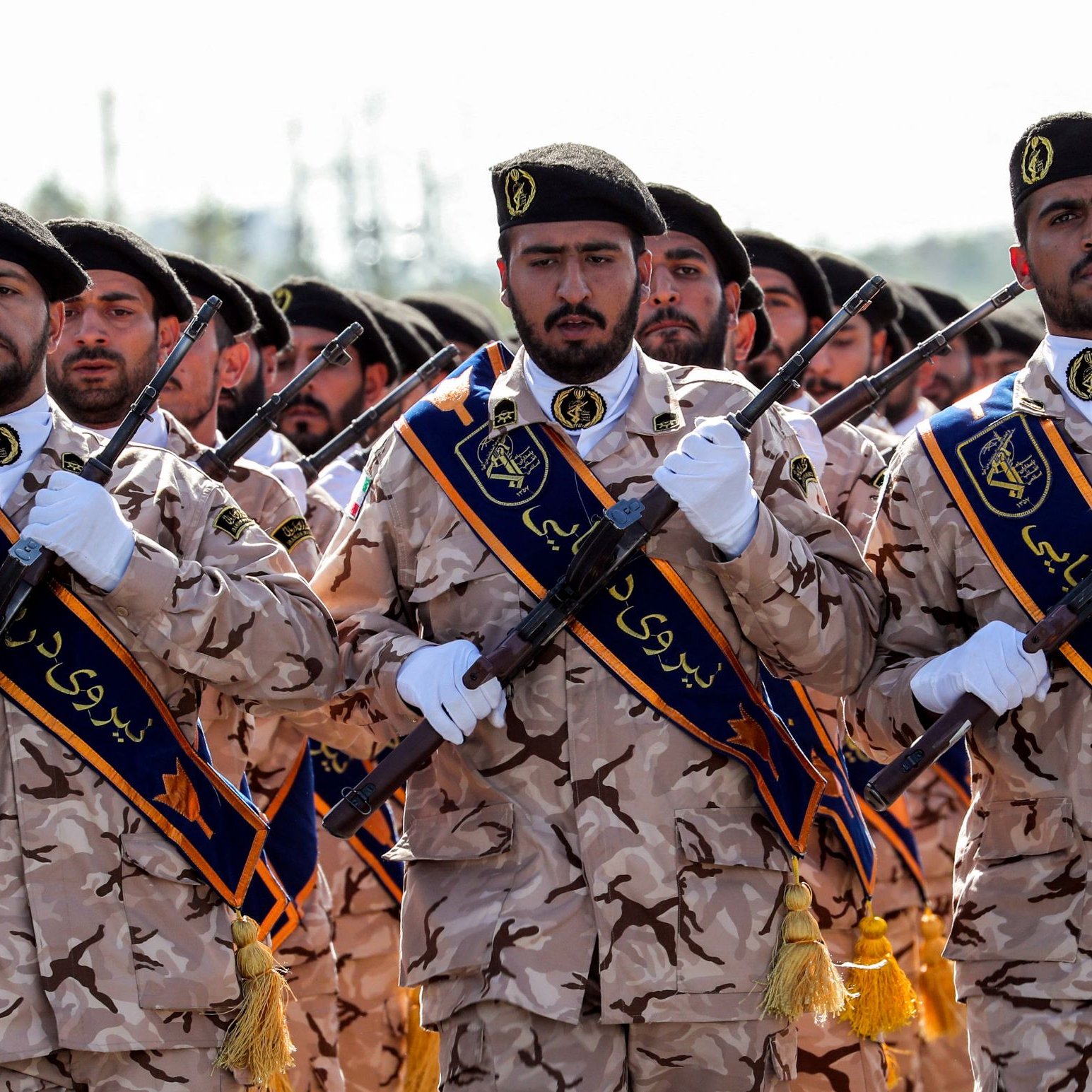 L'image montre un groupe de soldats en uniforme. Ils portent des uniformes camouflage et des képis distinctifs. Certains d'entre eux tiennent des fusils et affichent une attitude de discipline et de détermination. Des banderoles avec des inscriptions sont visibles sur leurs uniformes, et ils défilent en formation, visiblement lors d'une cérémonie ou d'un événement militaire. Le fond semble naturel, peut-être un terrain en extérieur.