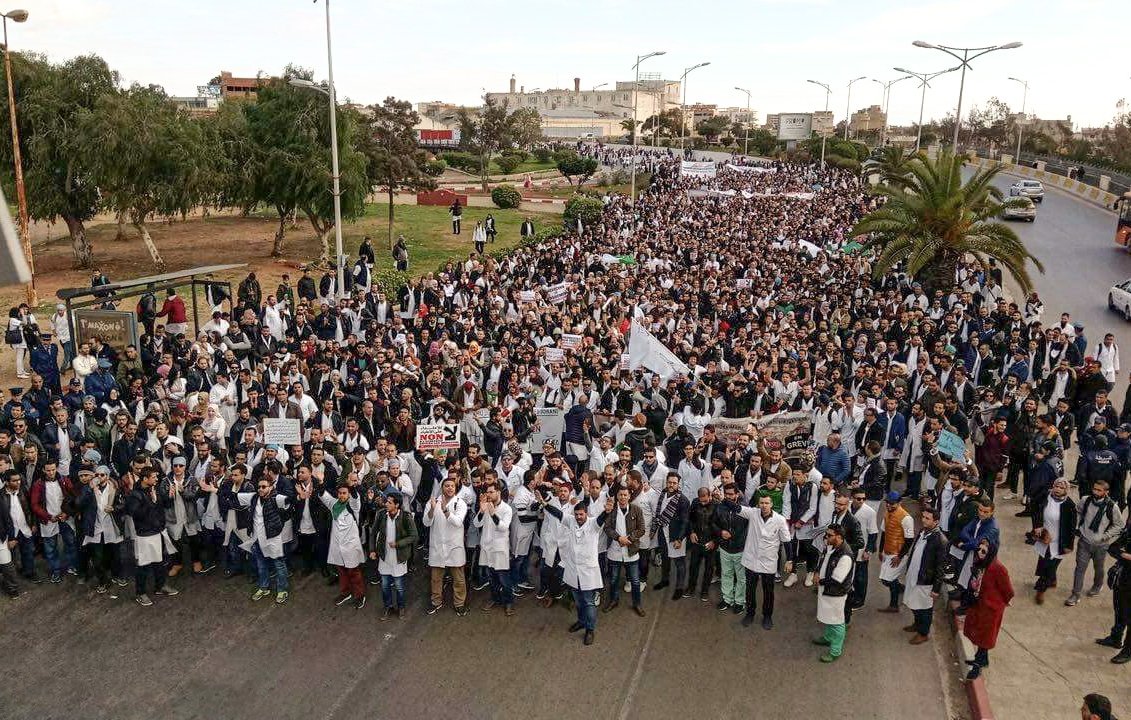 L'image montre une grande foule de manifestants rassemblés dans une rue, probablement pour une manifestation ou une protestation. Beaucoup de participants portent des blouses blanches, ce qui pourrait indiquer qu'ils sont des professionnels de la santé ou des étudiants en médecine. Ils agitent des panneaux et des drapeaux, exprimant leurs revendications. L'environnement semble urbain, avec des arbres et des bâtiments en arrière-plan. L'ambiance générale paraît mobilisée et solidaire.