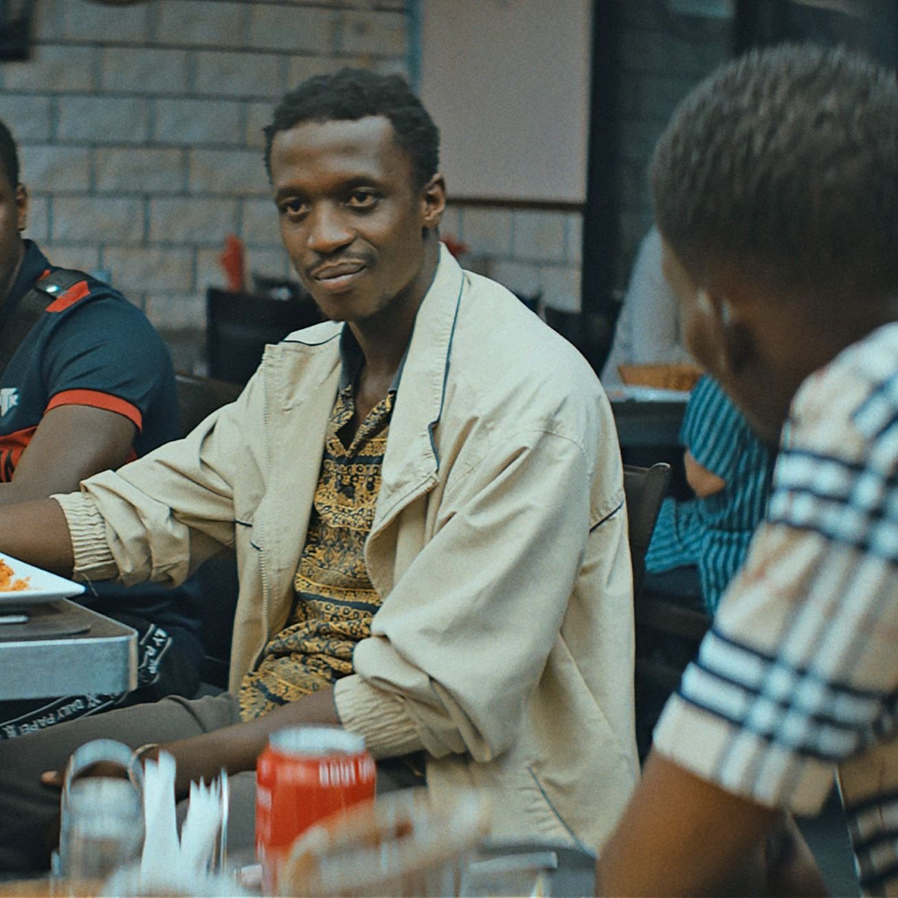 L'image montre un groupe de personnes assises dans un restaurant. Au premier plan, un homme souriant, portant une chemise claire et un motif coloré, semble engager une conversation. Deux autres hommes sont également visibles : l'un avec un maillot noir et rouge, et l'autre portant une chemise à carreaux. Les tables sont garnies de plats de nourriture, et l'ambiance est détendue, typique d'un moment convivial entre amis.