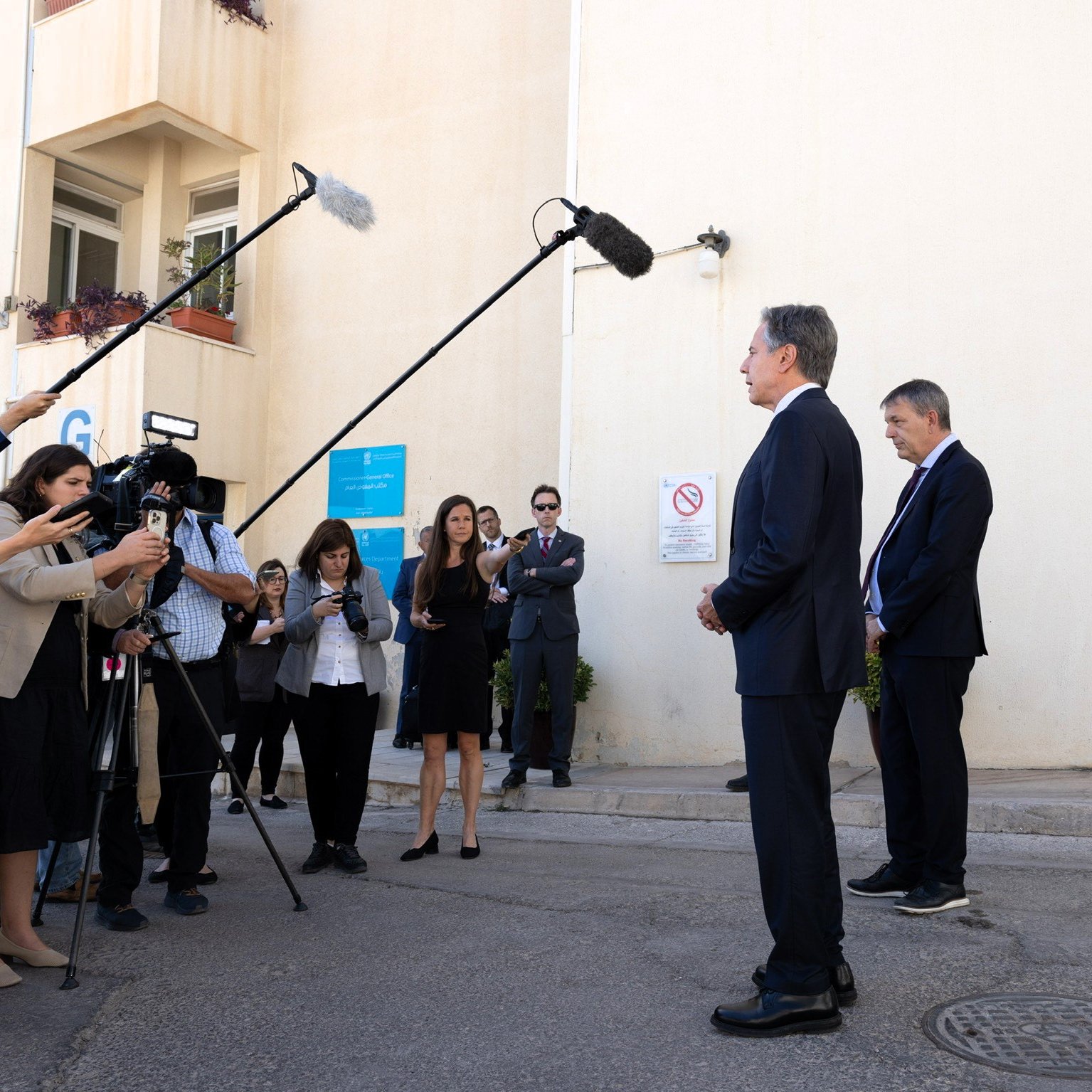 Nell'immagine si può vedere una scena di una conferenza stampa all'aperto. Due persone, vestite in abiti eleganti, sono in piedi di fronte a un muro chiaro, mentre un gruppo di giornalisti le circonda. Alcuni giornalisti stanno utilizzando microfoni e telecamere per registrare o fare domande. Dietro di loro, ci sono alcuni cartelli e avvisi su un edificio. L'atmosfera sembra formale e professionale, con un focus sulle dichiarazioni dei due individui in primo piano.