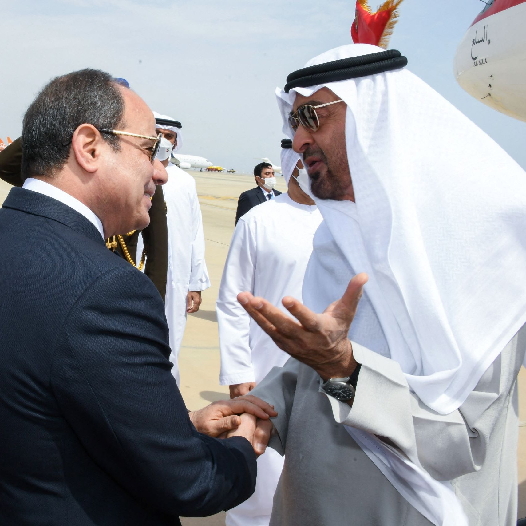 L'image montre une rencontre entre deux dirigeants. Ils semblent être en conversation amicale, avec des gestes visibles pour accompagner leurs paroles. L'un des deux est vêtu d'un costume formel, tandis que l'autre porte des vêtements traditionnels arabes, incluant un keffieh. En arrière-plan, on peut apercevoir des membres du personnel et des avions, suggérant que la scène se déroule dans un aéroport. L'atmosphère paraît conviviale et respectueuse.