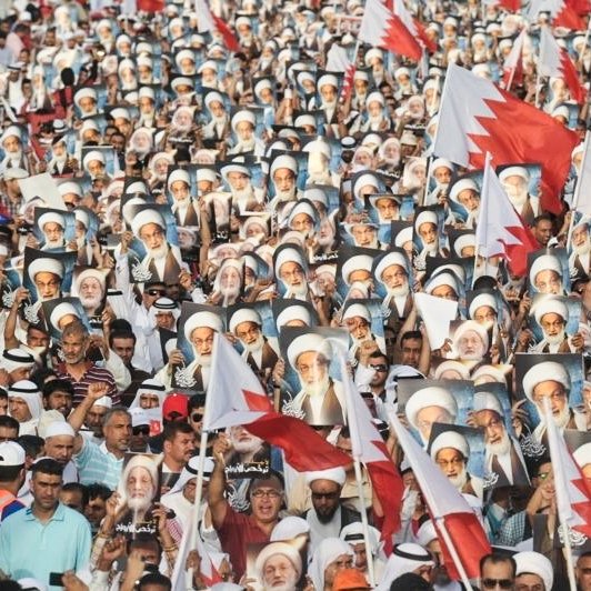 الصورة تُظهر تجمعاً حاشداً لمتظاهرين في البحرين. المشاركون يحملون أعلام البحرين، بالإضافة إلى صور لشخصيات معروفة. الجو يبدو حماسياً، حيث يرتدي العديد من المشاركين الثياب التقليدية. المناظر تعكس روح التضامن والمشاركة الفاعلة في الحدث.