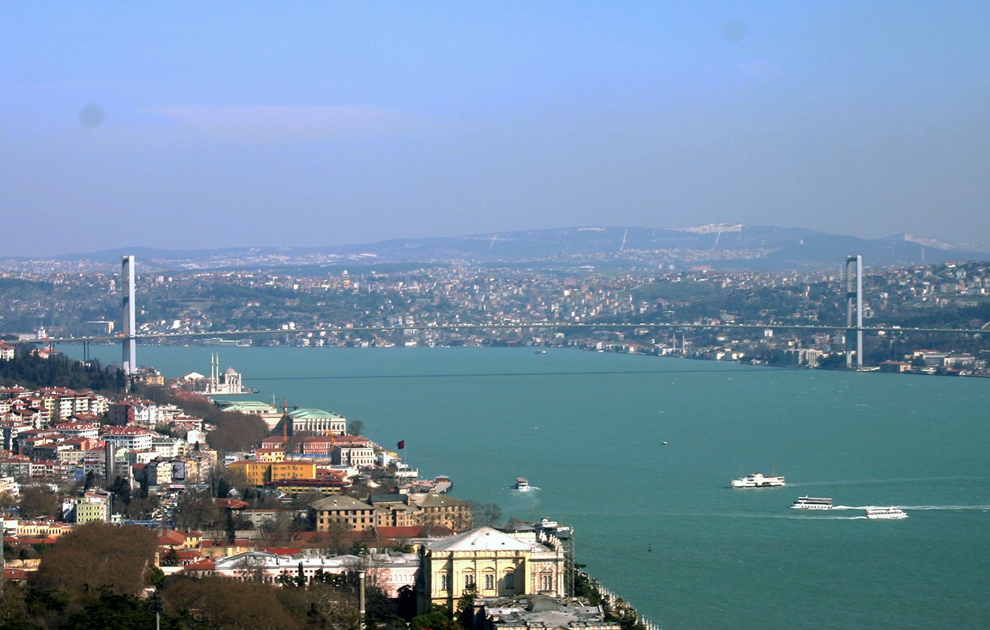 L'image montre un panorama d'une ville au bord d'une grande étendue d'eau. On aperçoit un pont moderne qui traverse la rivière, reliant les deux rives. À gauche, des bâtiments s'échelonnent sur la colline, tandis qu'à droite, on peut voir des constructions historiques. Le ciel est clair, et l'ensemble du paysage évoque une atmosphère tranquille et pittoresque. On distingue également des bateaux naviguant sur la rivière.