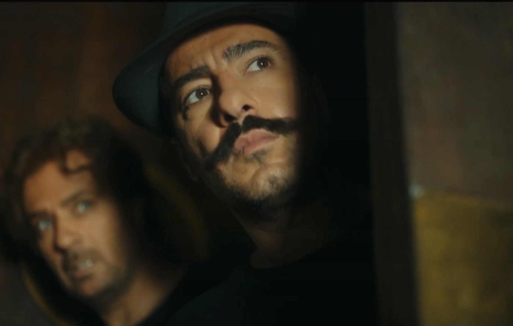 در این تصویر، دو مرد در یک محیط تاریک و مرموز دیده می‌شوند. مردی در جلو دارای کلاهی سیاه و سبیلی خاص است که به جلو نگاه می‌کند و حالت تفکر یا نگرانی دارد. مرد دیگر در پس‌زمینه، با ظاهری نگران و معتمد، از سمت چپ او به تصویر نگاه می‌کند. نور به صورت جزئی روی صورت‌های آن‌ها می‌تابد و حس تنش یا خطر در صحنه وجود دارد.