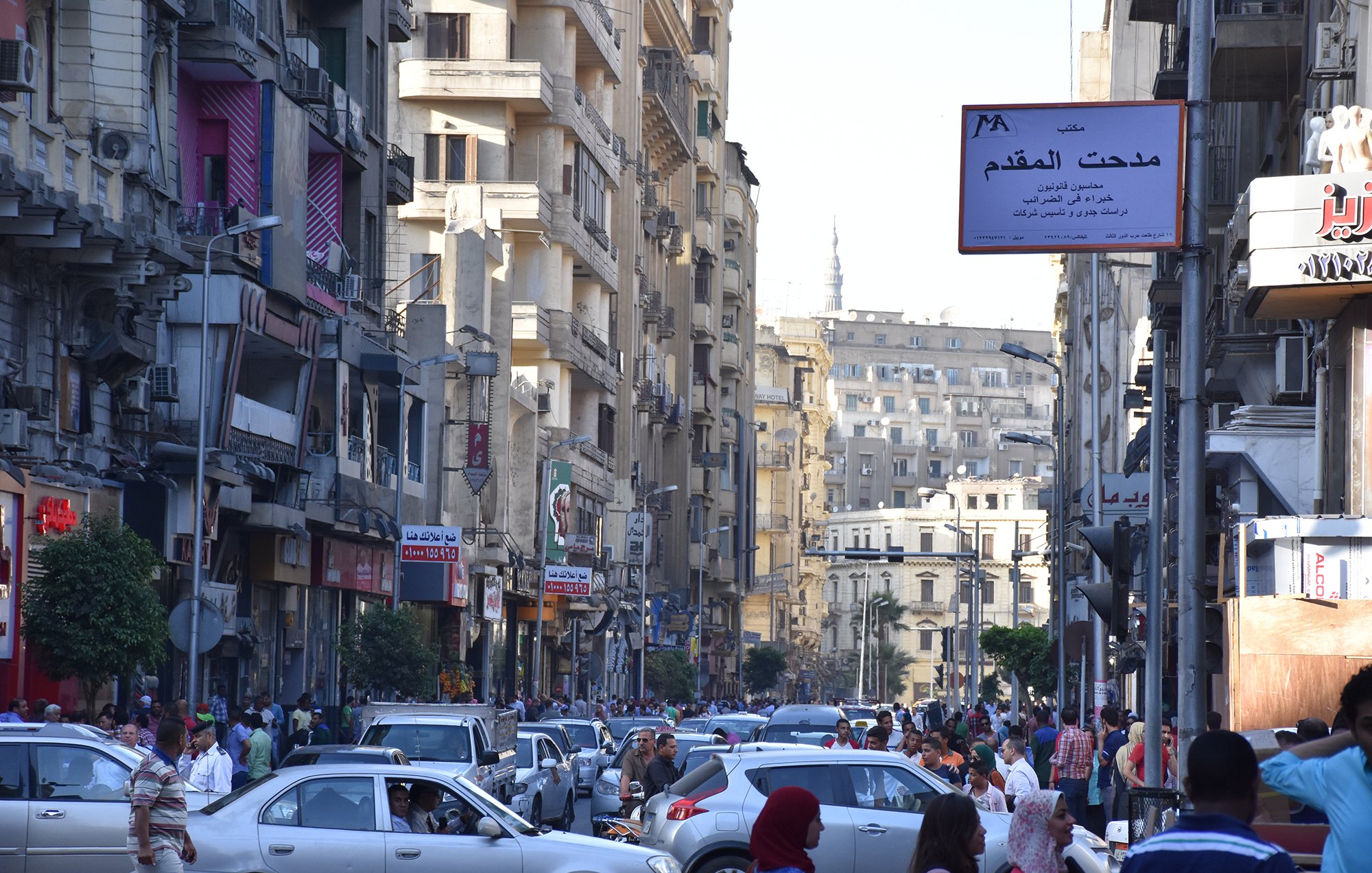 L'image montre une rue animée dans une ville, probablement une métropole. On peut voir de nombreux bâtiments de plusieurs étages qui bordent la route. Les voitures sont garées et circulent, créant un certain embouteillage. Une foule de personnes se déplace le long des trottoirs. Des panneaux de signalisation et des enseignes en arabe sont visibles, indiquant que cette scène se déroule dans un pays arabophone. L'atmosphère semble dynamique et vivante, typique d'une grande ville.