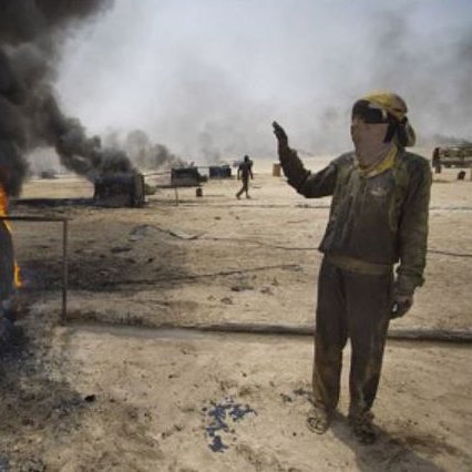 L'image montre un homme dans un environnement industrialisé, à proximité de réservoirs en feu. Des flammes et une épaisse fumée noire s'élèvent de plusieurs conteneurs. L'homme semble faire un geste d'alerte ou de signalisation, et il porte des vêtements de travail, suggérant qu'il se trouve dans une zone à risque, possiblement liée à l'exploitation pétrolière. En arrière-plan, d'autres personnes sont visibles, mais leur activité n'est pas clairement définie. L'atmosphère de la scène est sombre et montre les dangers liés aux incendies industriels.